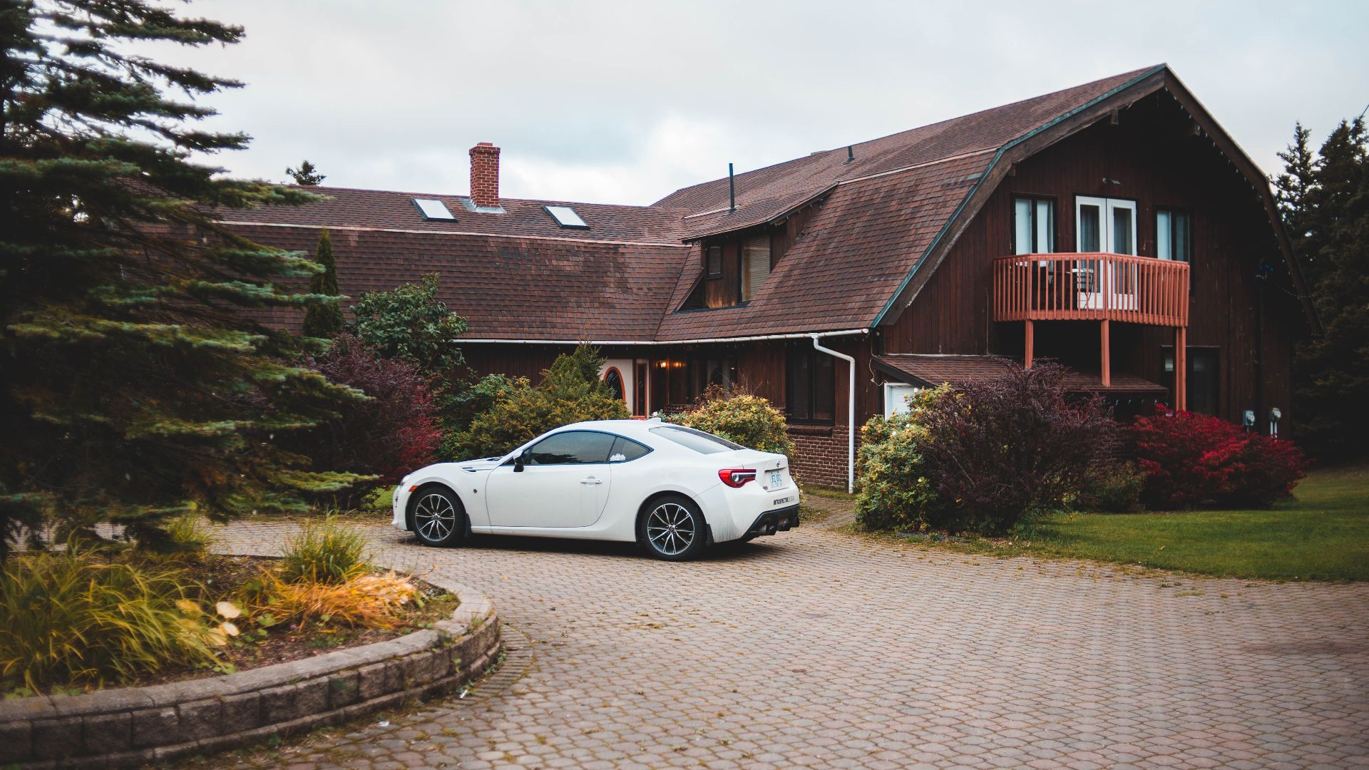 Rustic house with a white car parked on a cobblestone driveway amidst lush greenery.