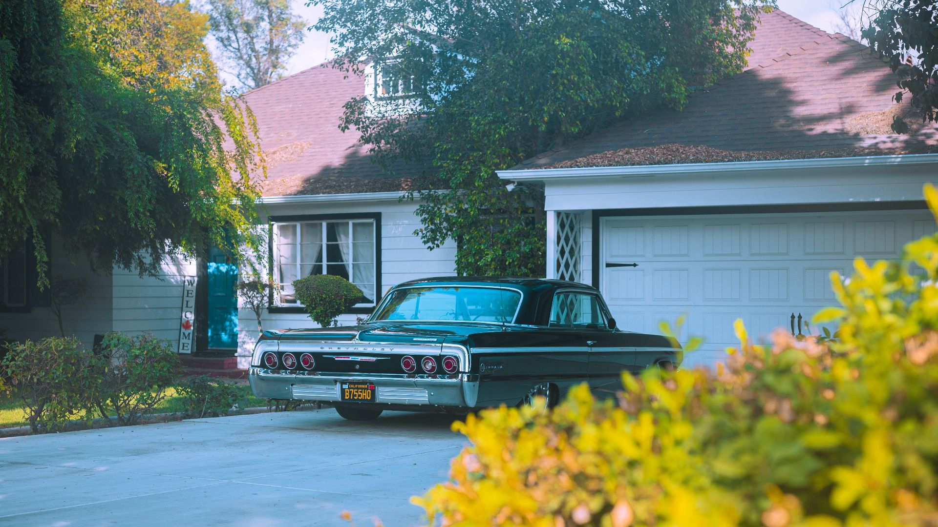 A classic black Chevrolet Impala parked in a suburban driveway surrounded by greenery.
