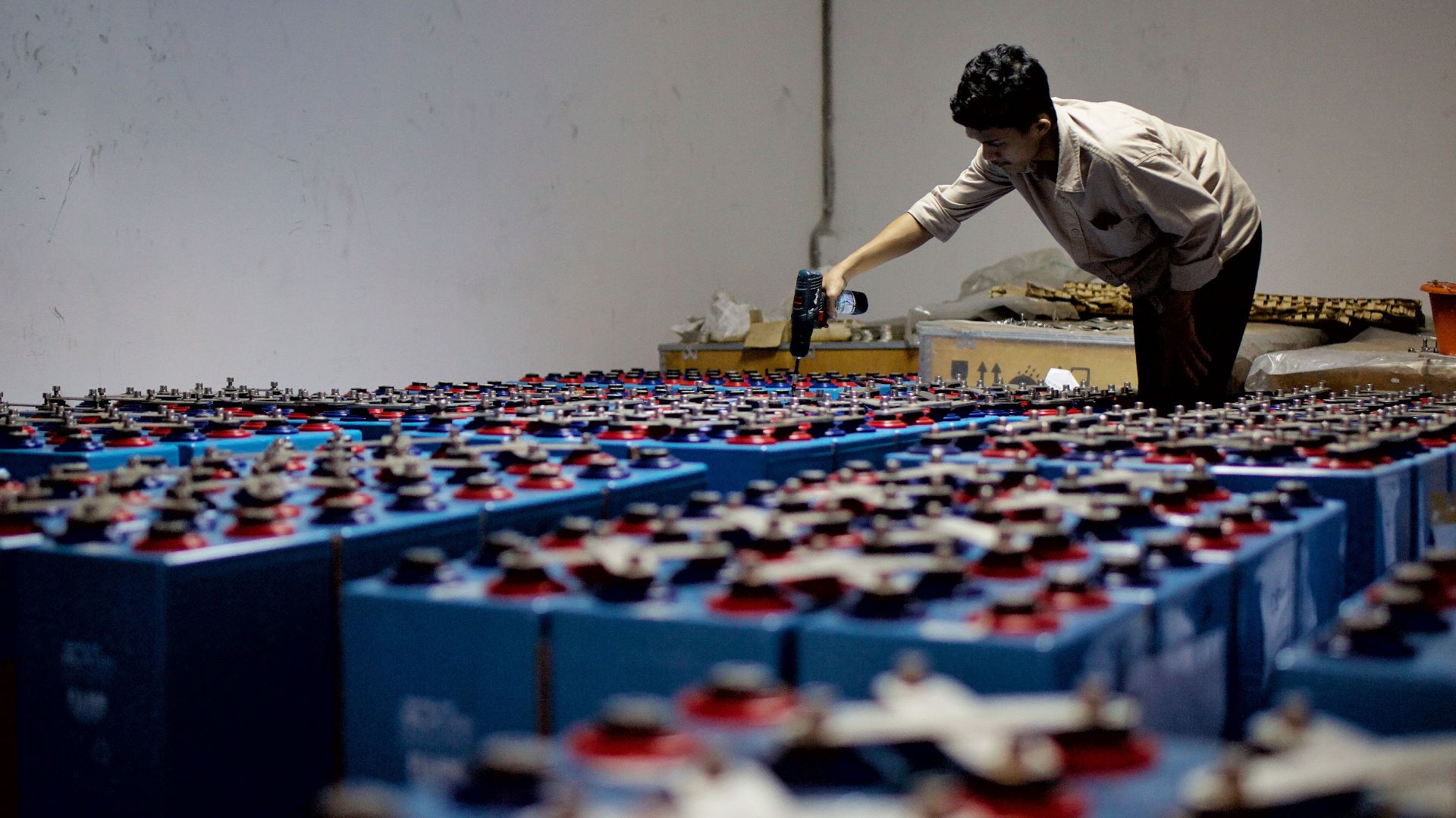 A worker checking many industrial batteries inside a facility. Indoor, industrial setting.