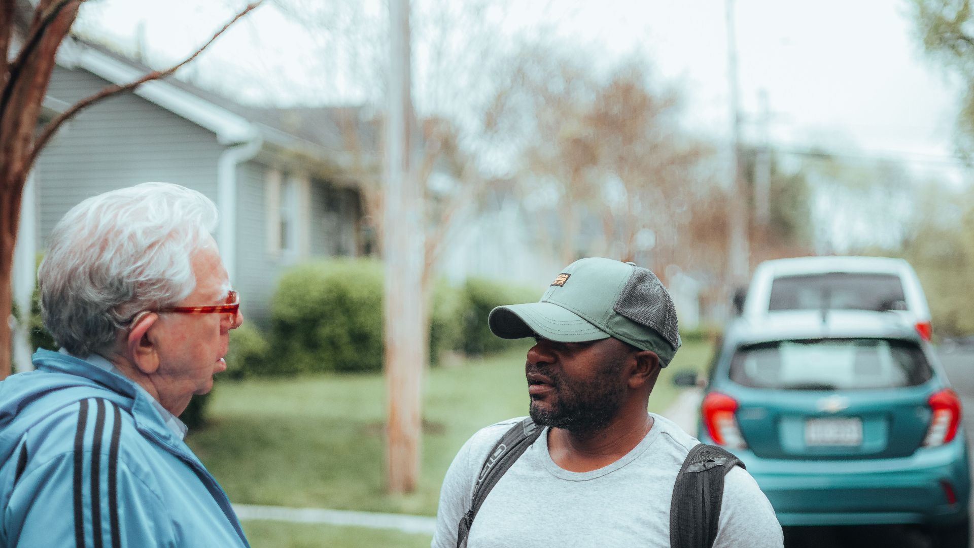 Two men engaged in conversation on a suburban sidewalk with parked cars.