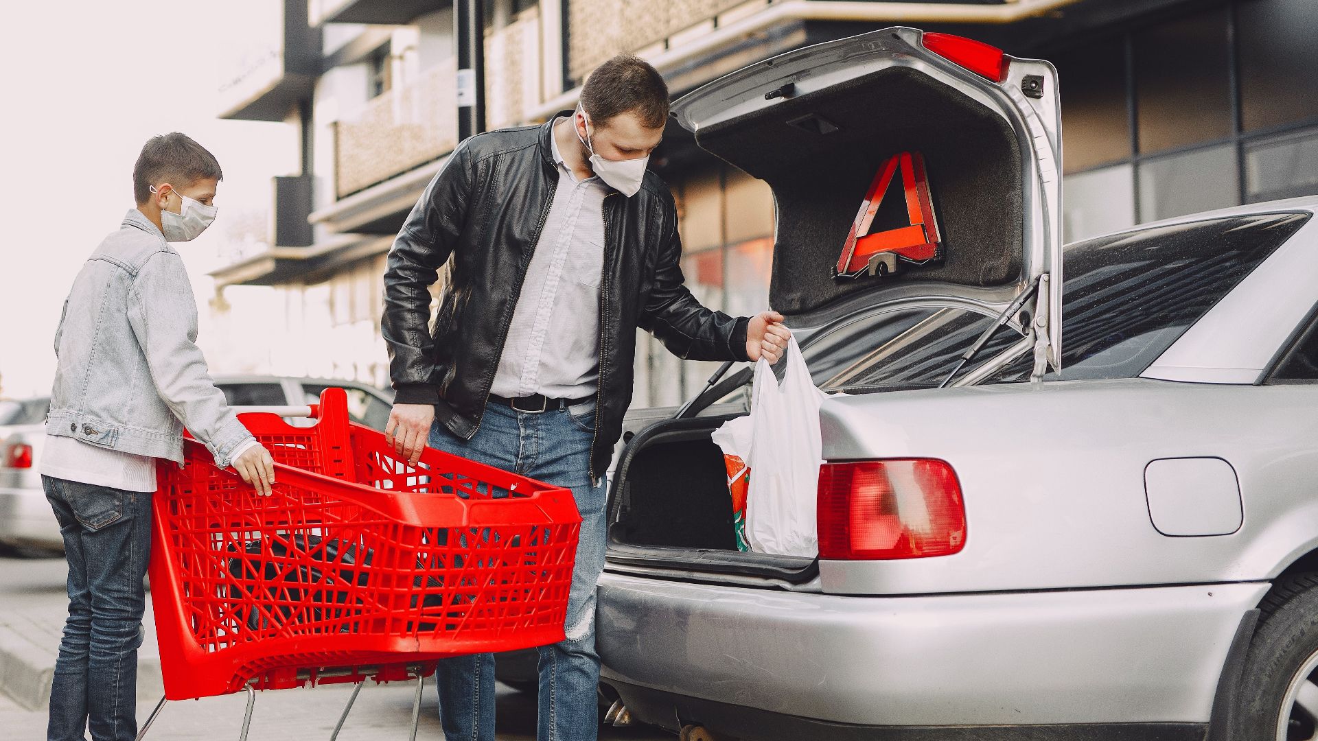 Young father and little son in casual clothes and protective masks moving shopping bags from supermarket trolley to trunk modern building parking