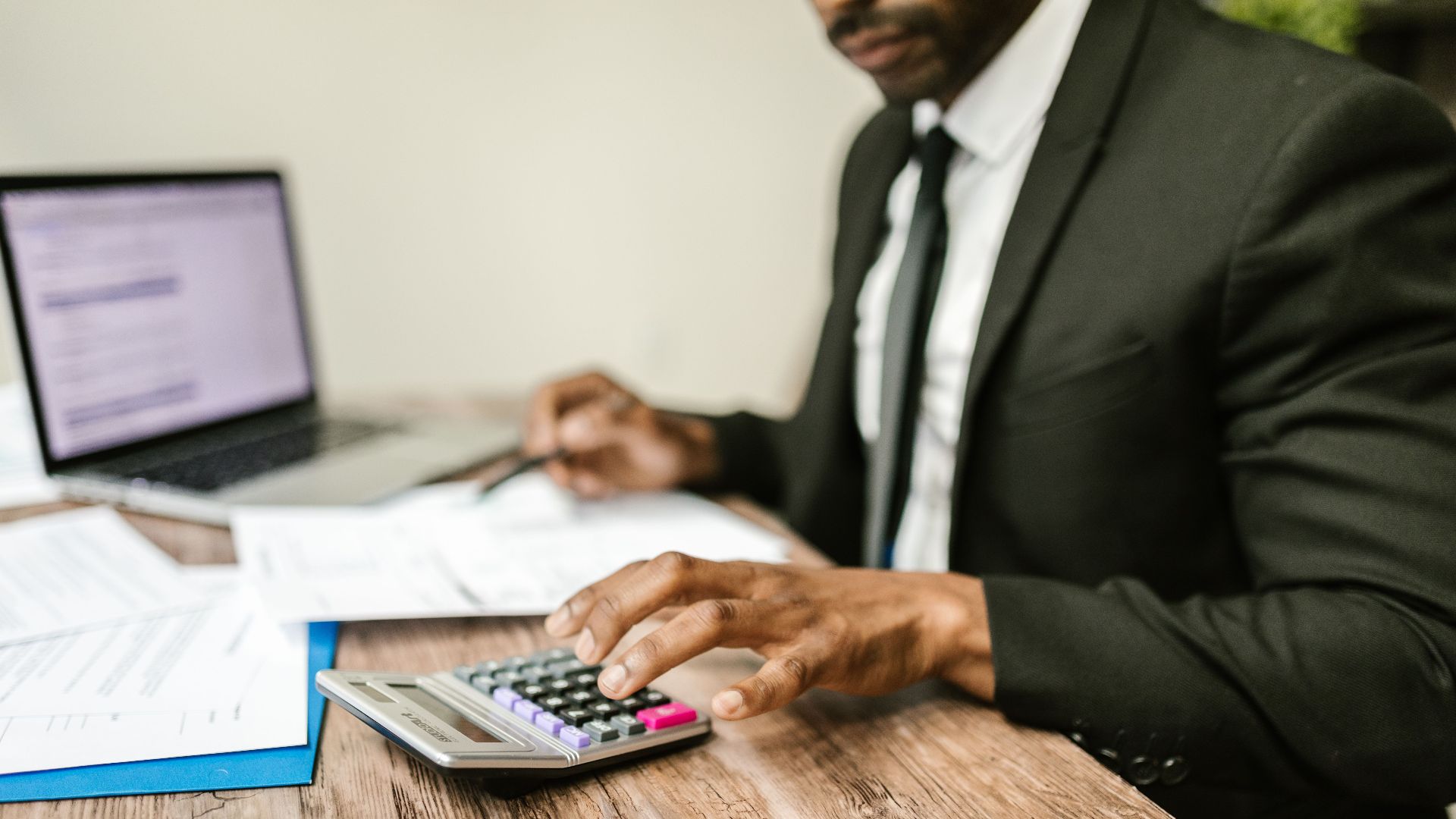 A businessman in a black suit using a calculator at a desk with financial documents and a laptop.