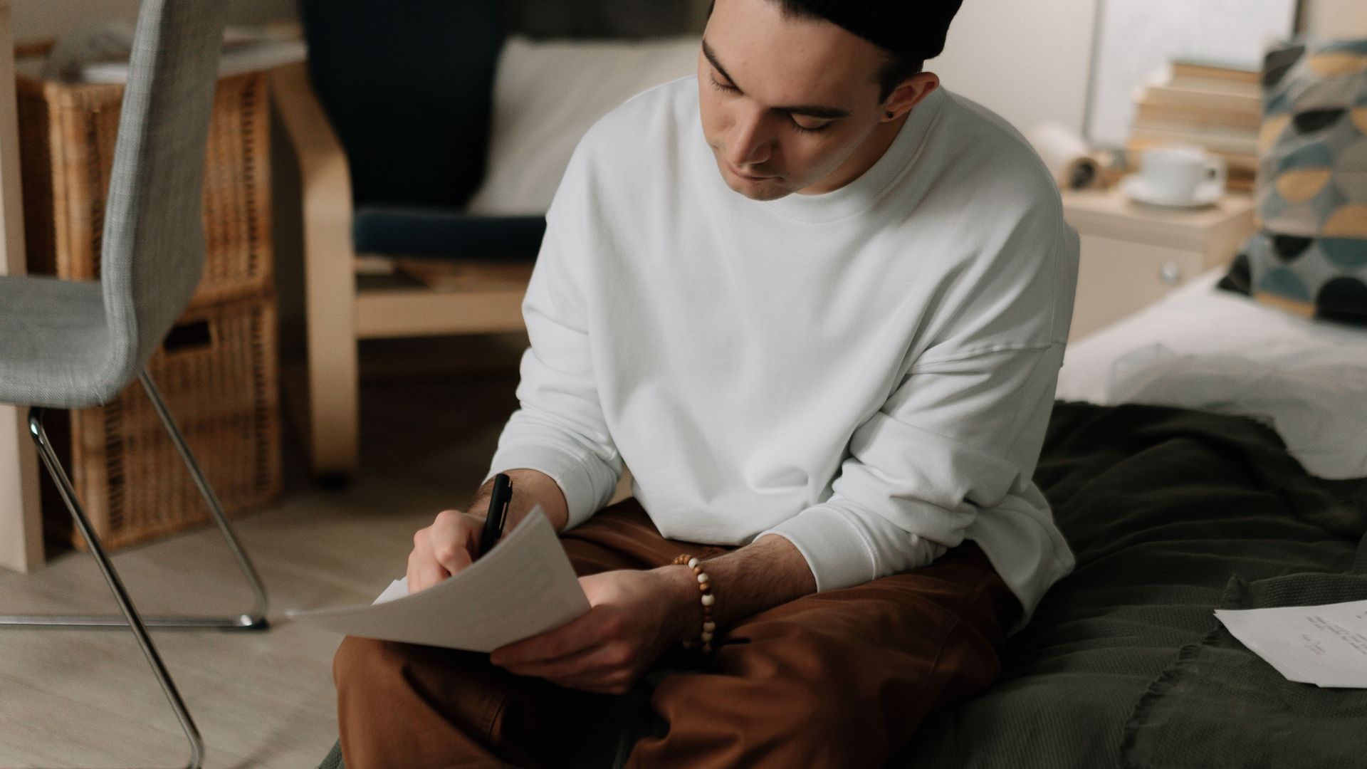 A man sitting on a bed writing in a notebook, wearing white long sleeves in a cozy room.