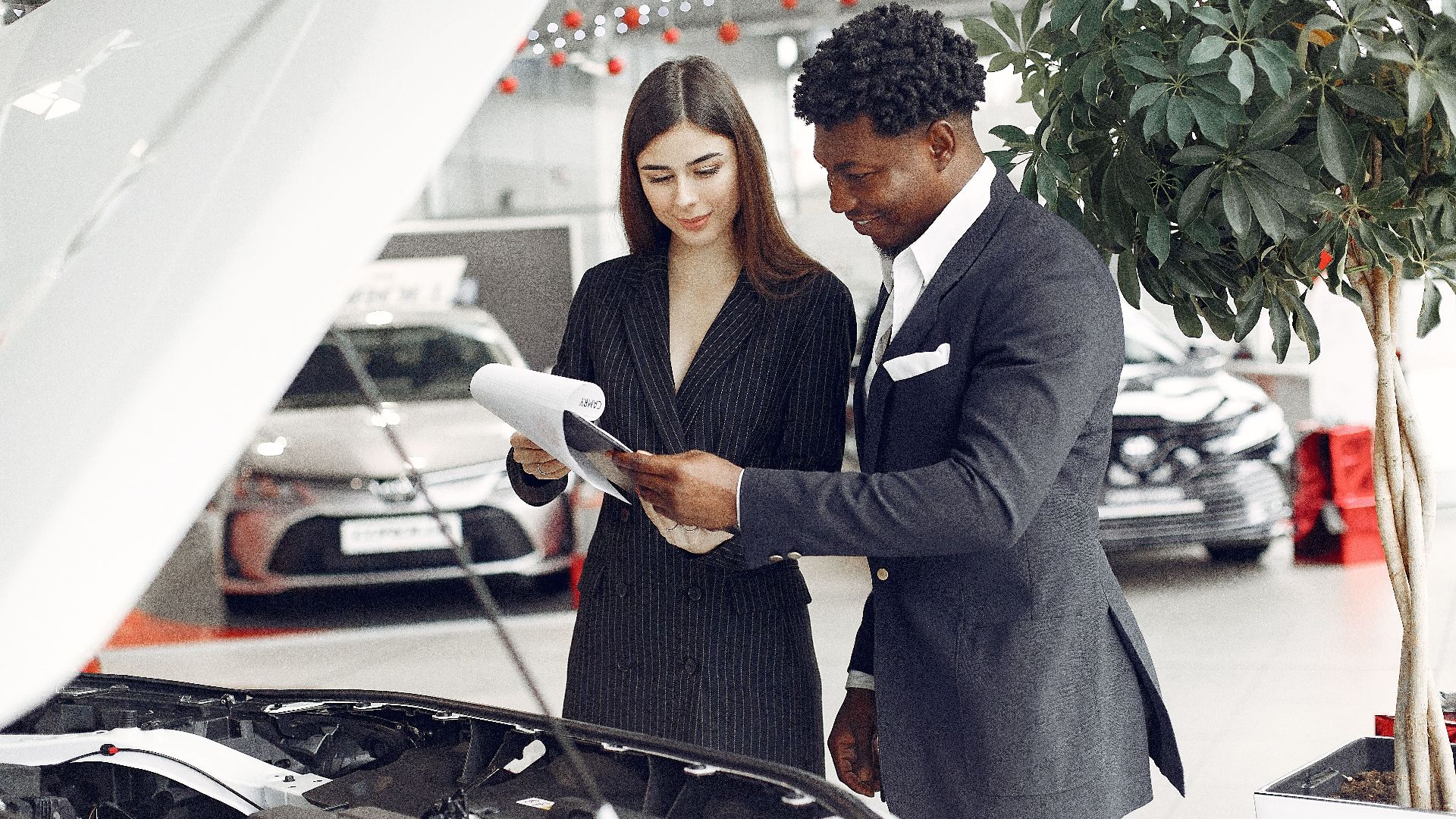 Confident smiling successful black businessman in classy formal suit standing near open motor hood in car showroom and discussing car characteristics with gorgeous female dealer