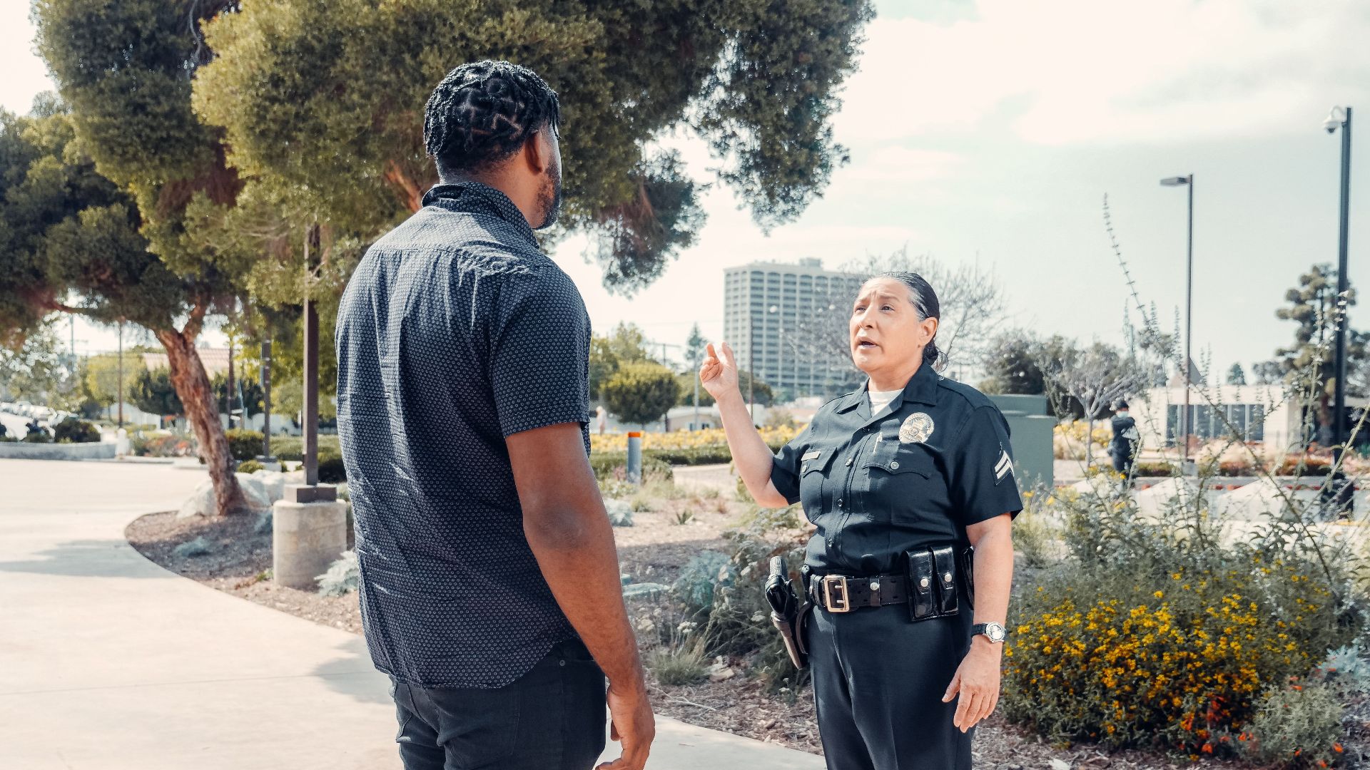 A police officer engages in conversation with a civilian in a park setting on a sunny day.