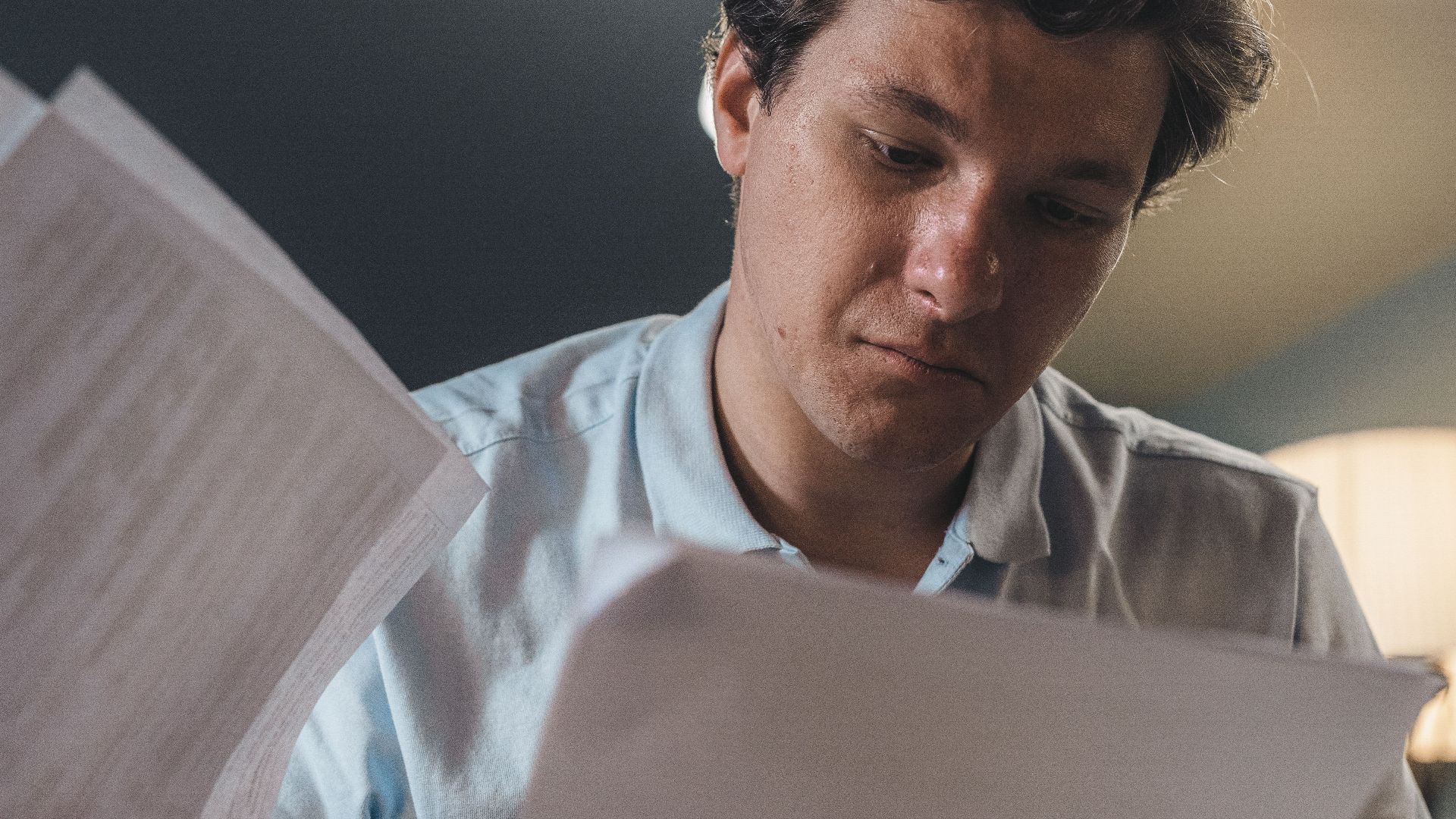 Serious man wearing a light blue shirt intently reading printed papers indoors under warm lighting.