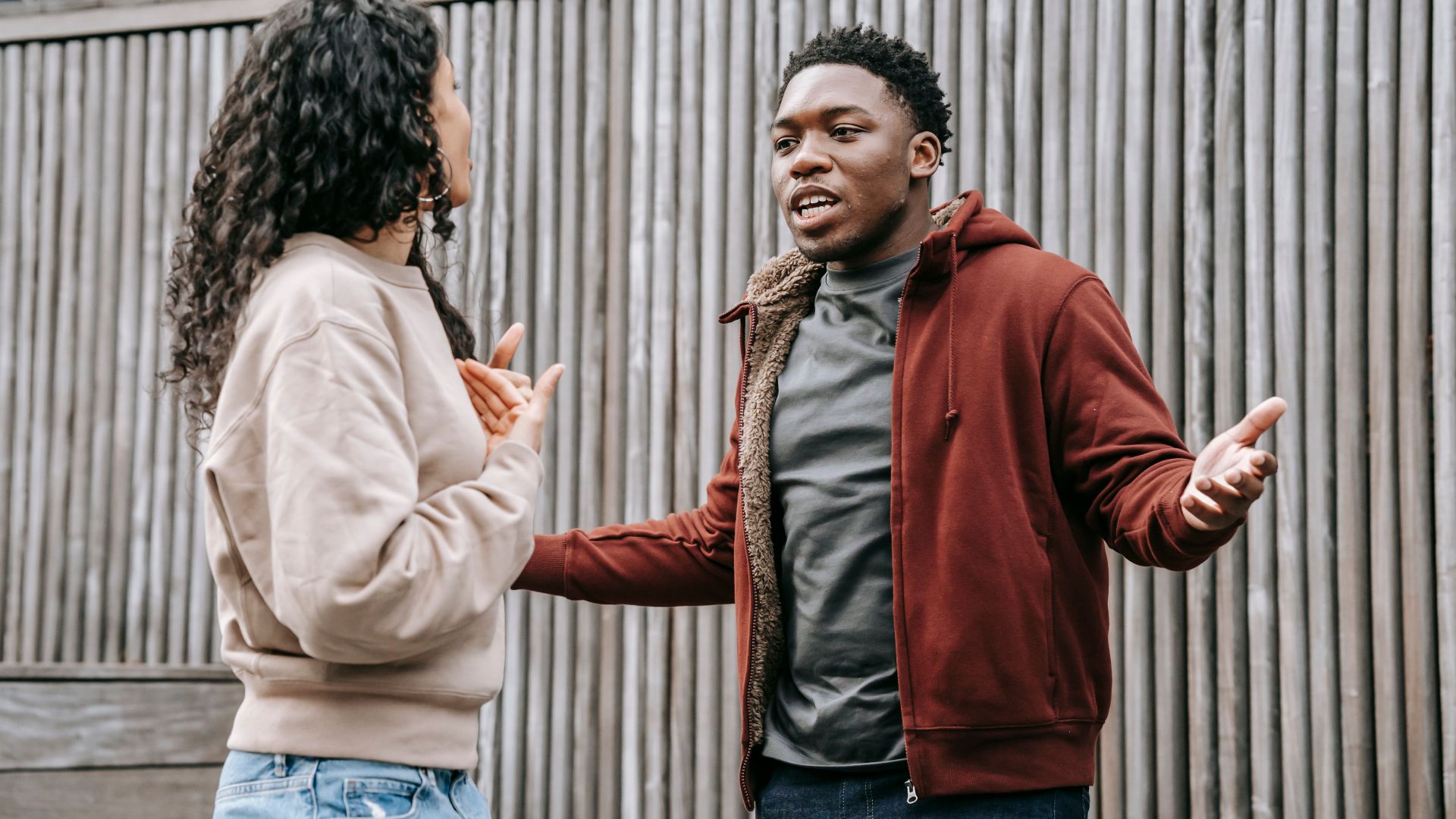 Angry African American boyfriend and ethnic girlfriend looking at each other while having emotional dispute on street near wooden fence