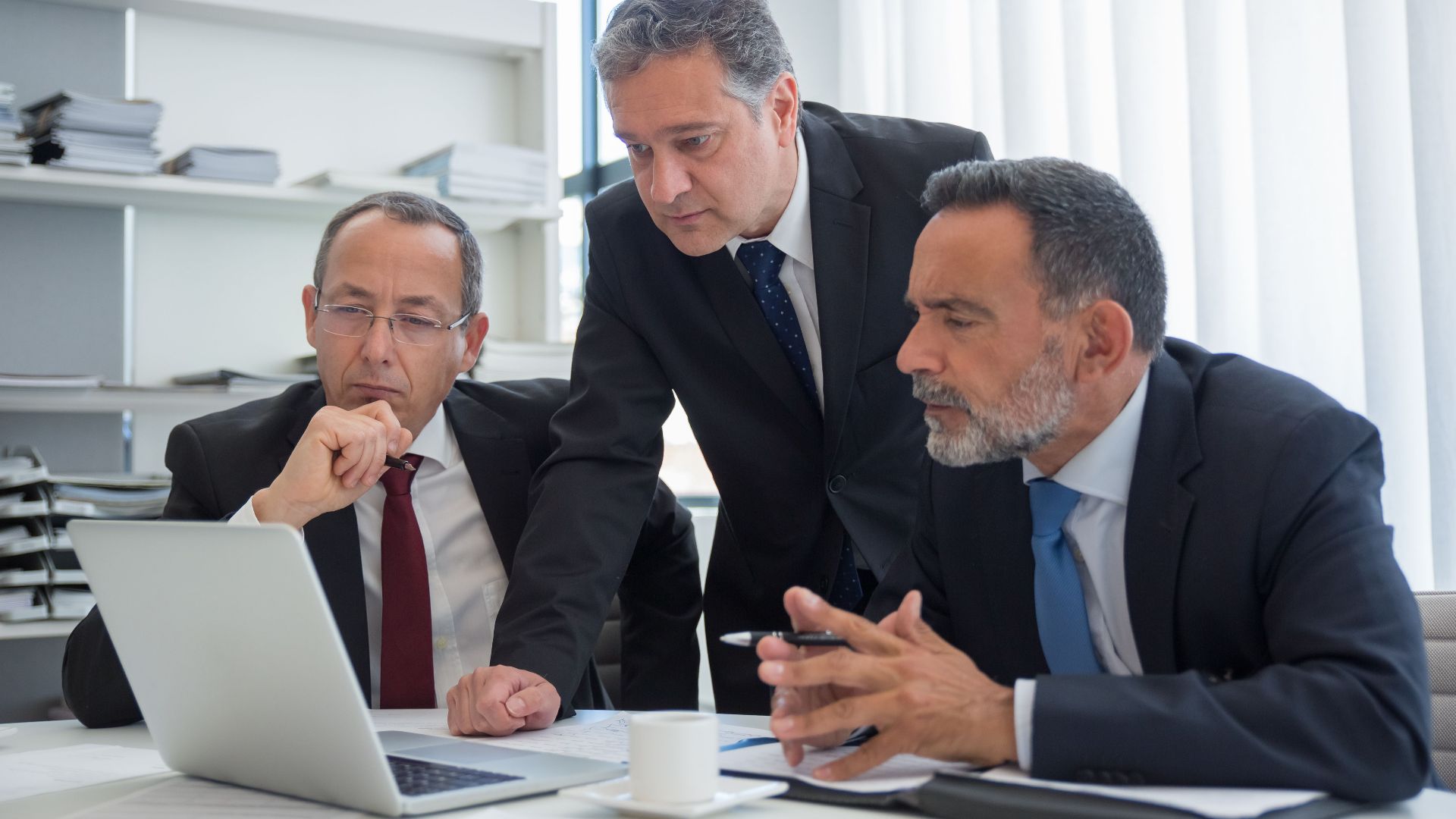 Three businessmen in suits collaborating in a modern office setting, focused on a laptop.