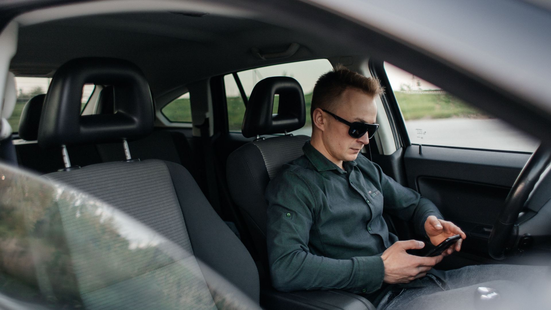 Young man using smartphone inside a parked car, wearing sunglasses.