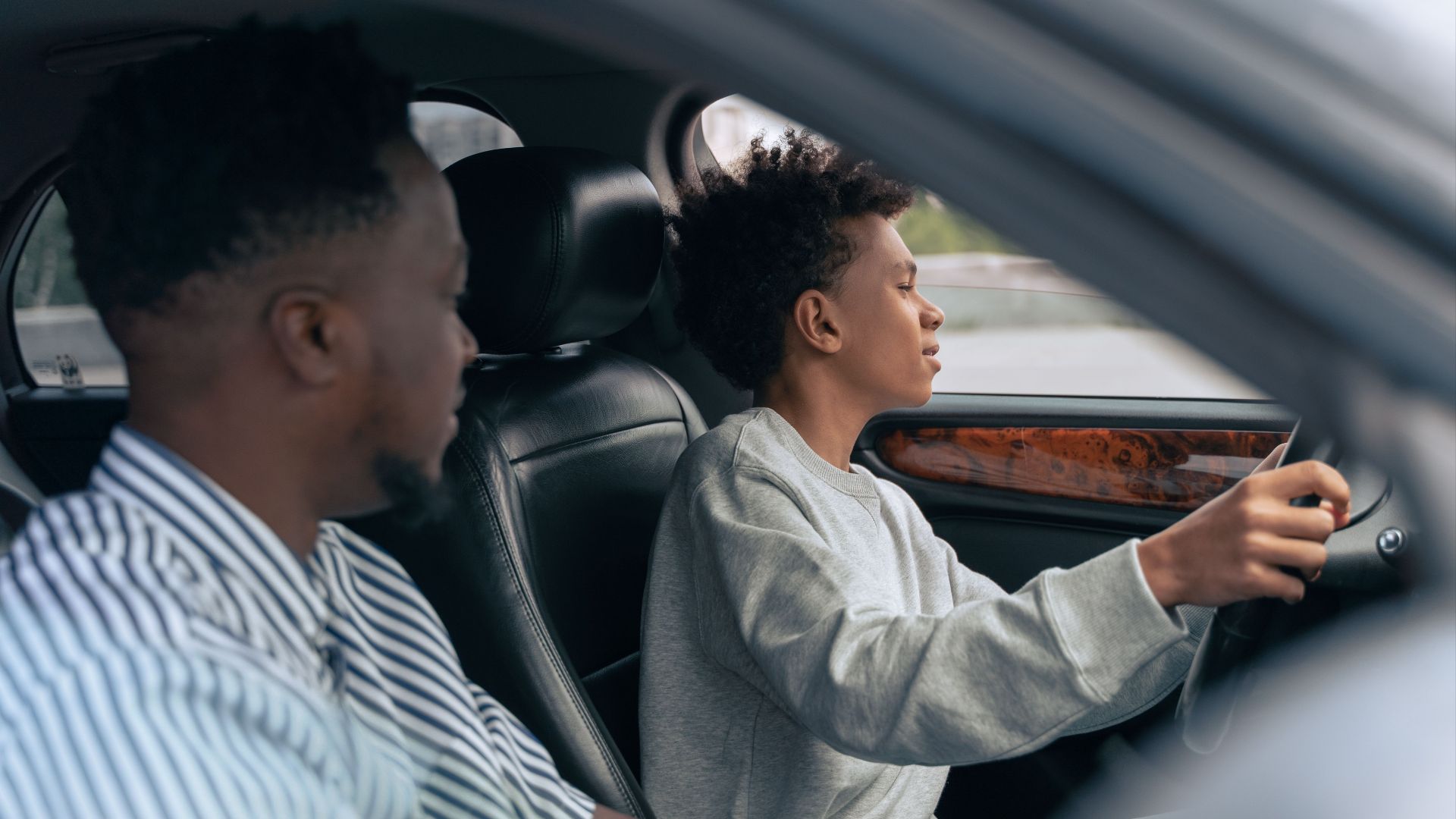 A father teaches his son to drive, sitting together in a car and discussing techniques. Side view indoors.