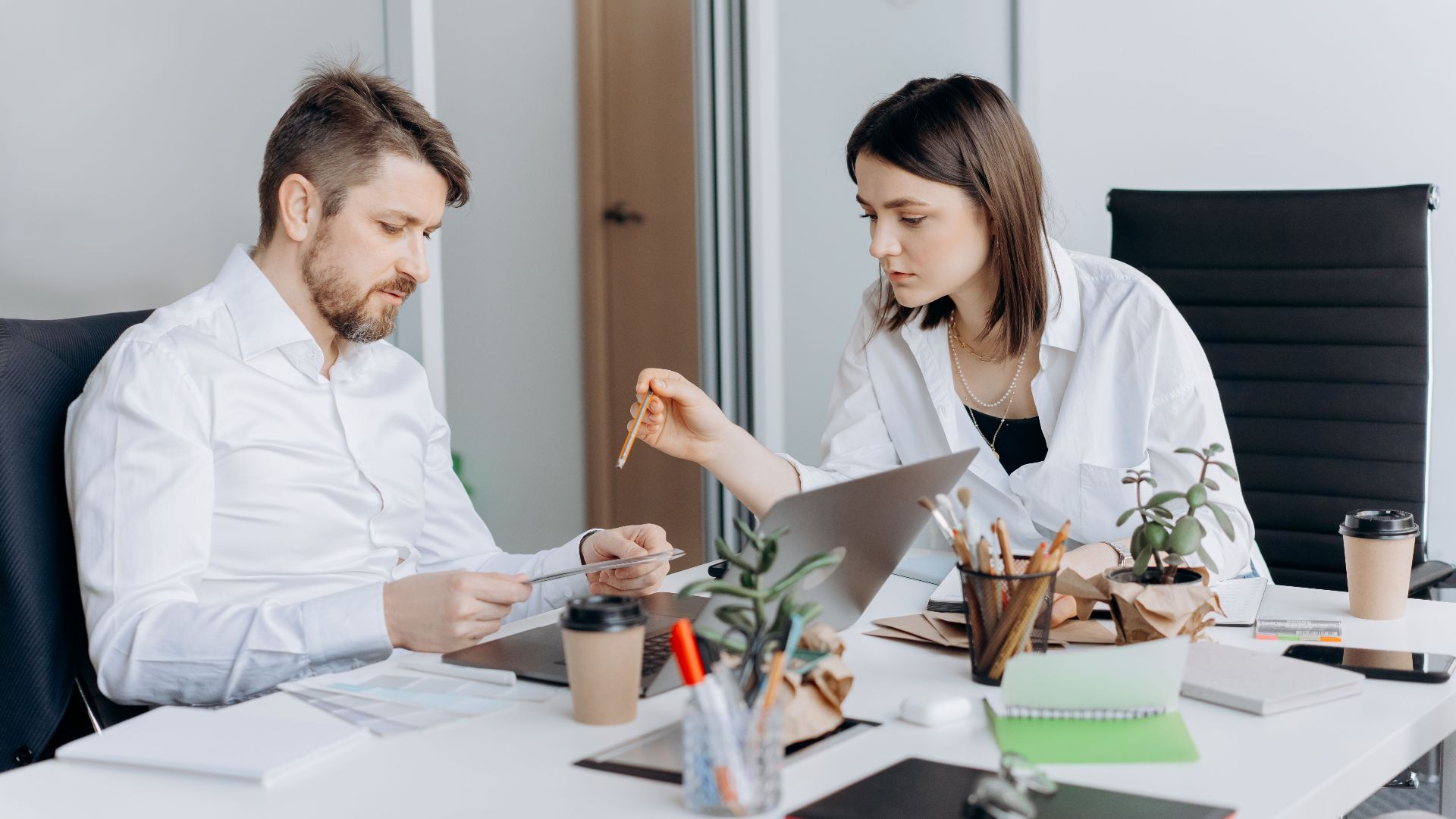 Two colleagues discussing work at a desk with laptops and coffee in a modern office setting.