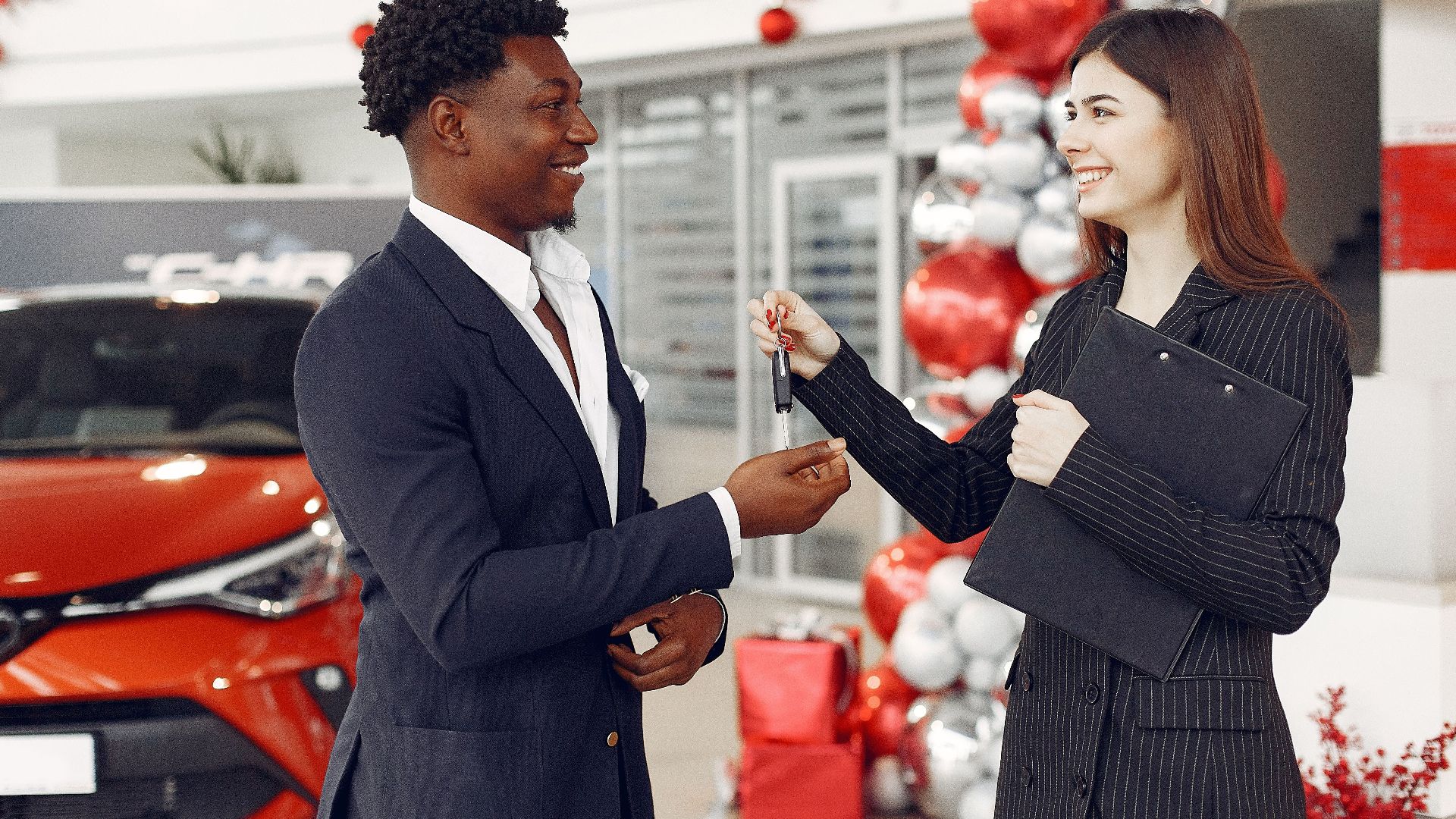 Side view of happy positive female dealer in formal classy wear passing car keys to  excited African American customer in trendy suit in car showroom