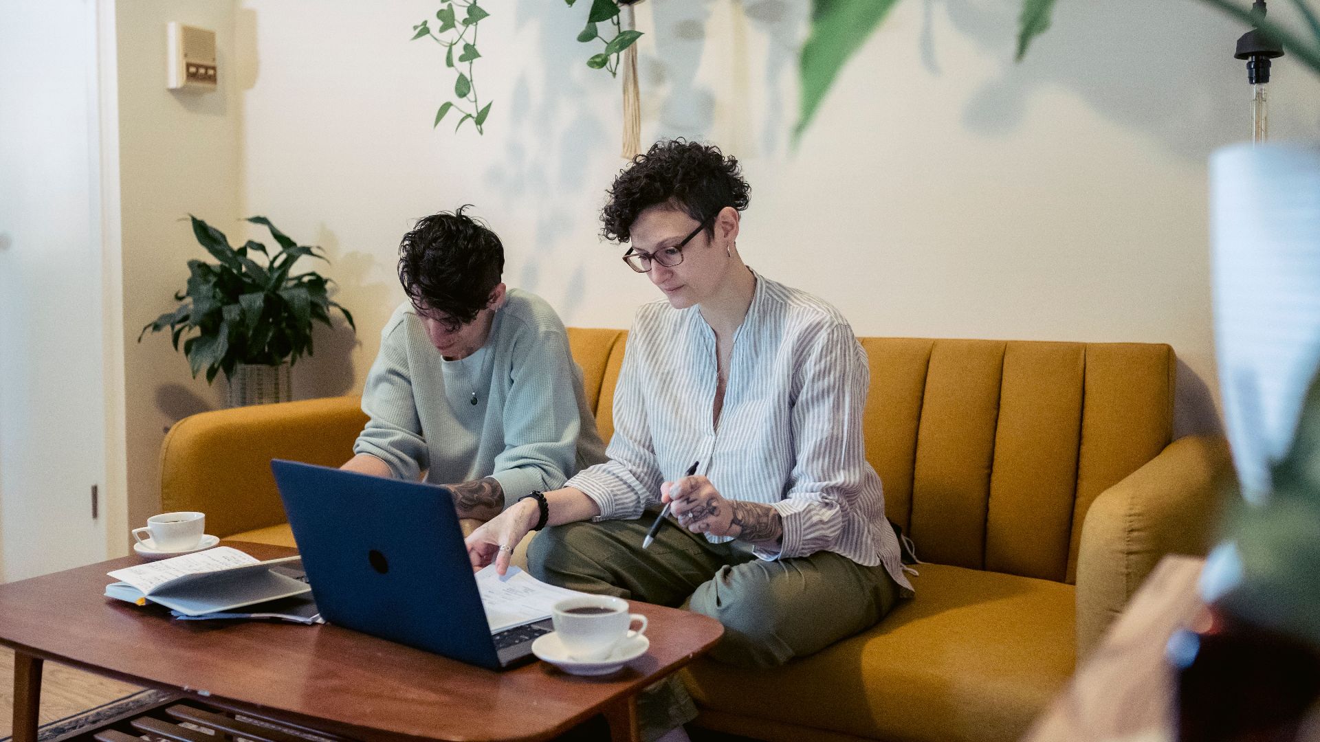 Concentrated young tattooed female freelancers with short dark hair in casual clothes sitting on comfortable sofa and working remotely on laptop