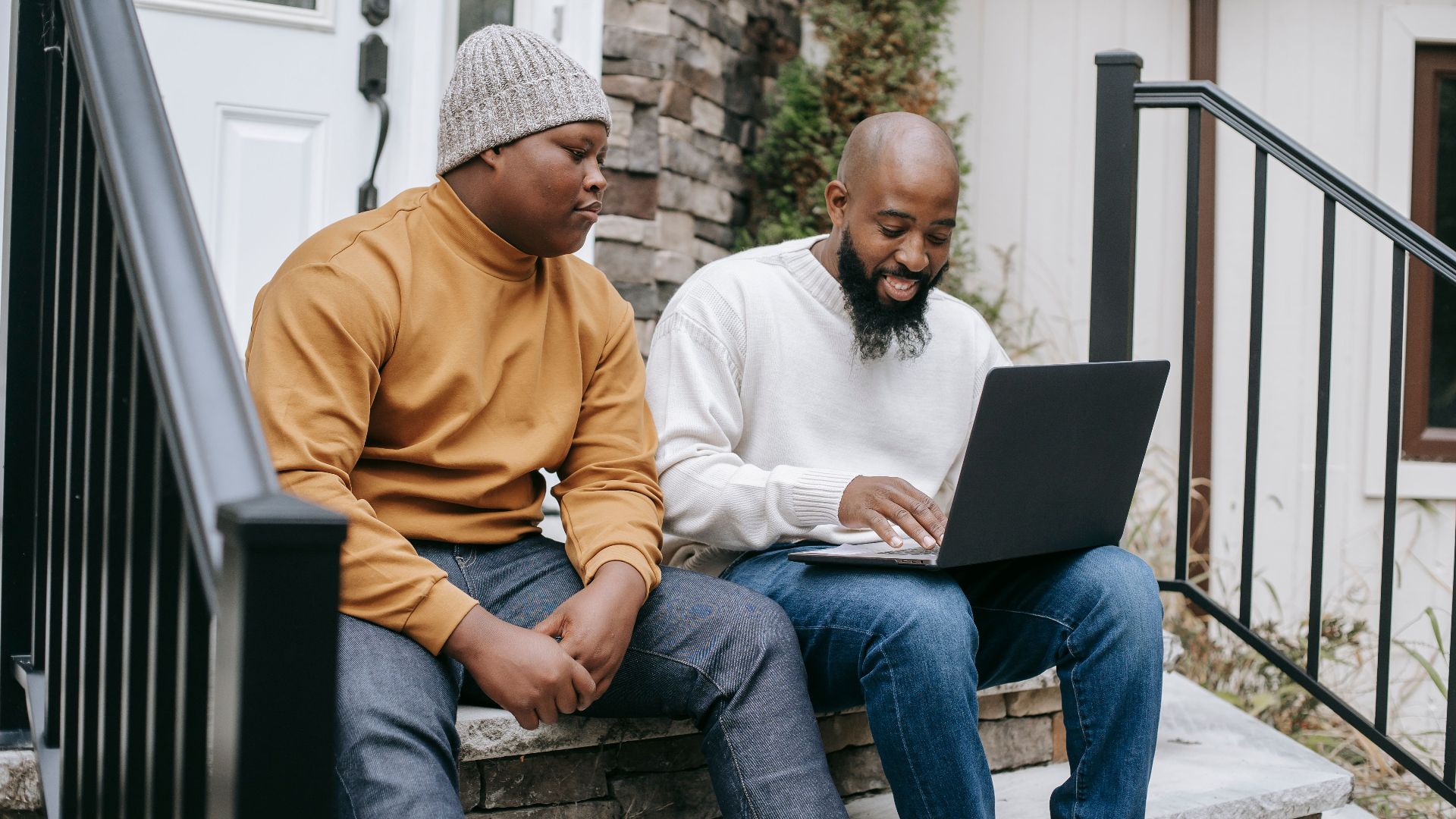 Positive bearded African American male remote worker sitting on stairs and typing on netbook keyboard with teen son