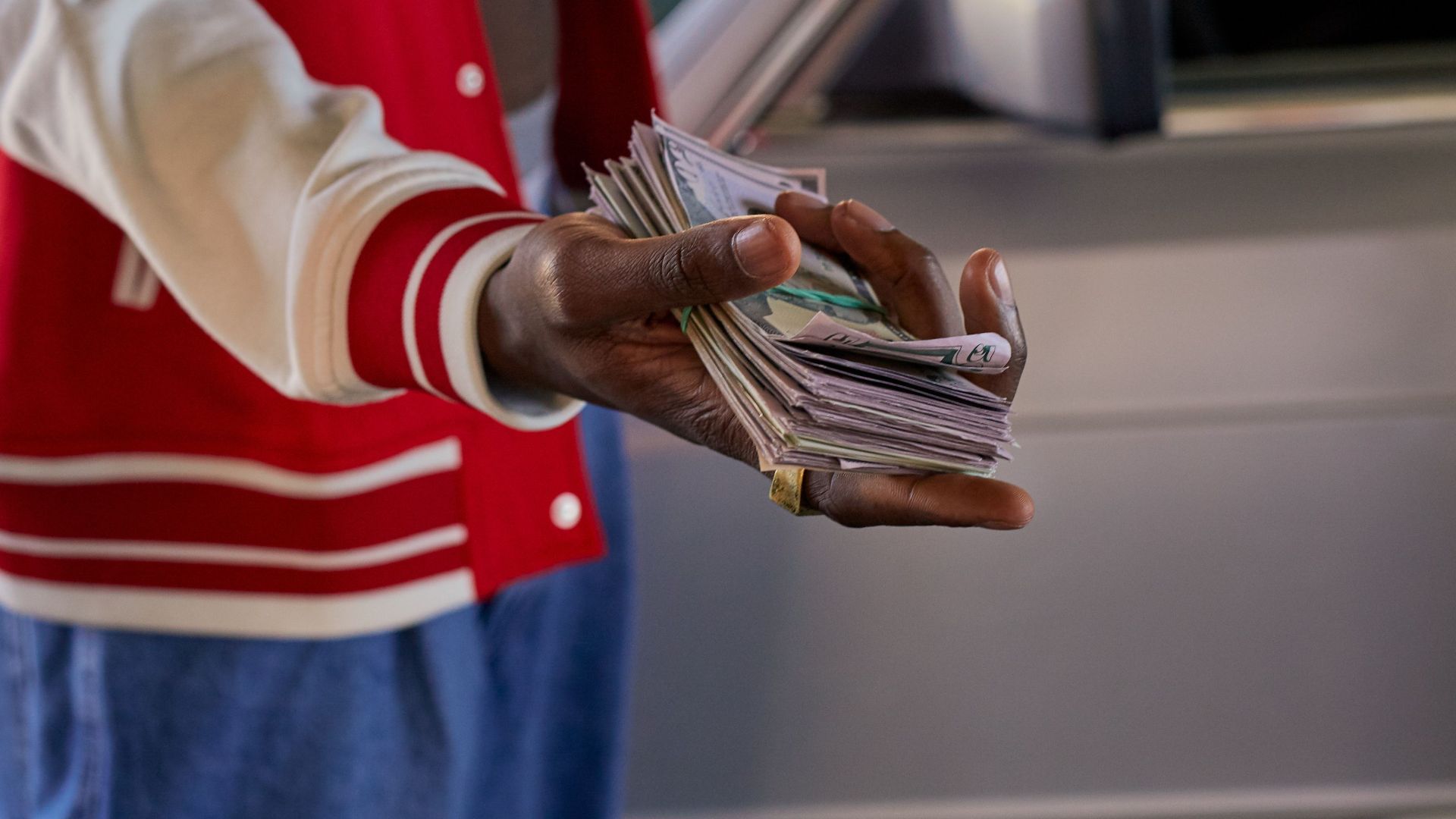 Close-up of a person offering a stack of cash in front of a car, symbolizes financial transaction.