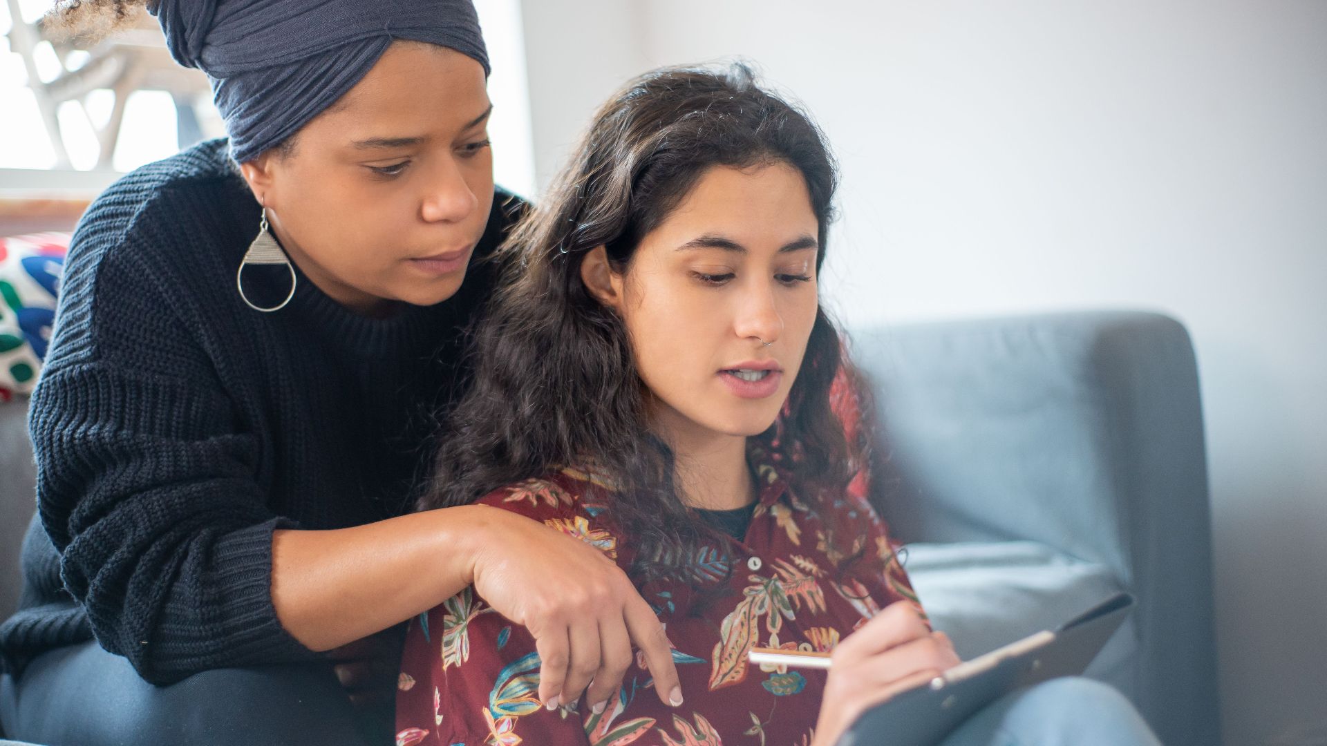 Two women sitting on a sofa sharing a thoughtful moment, one writing or drawing.