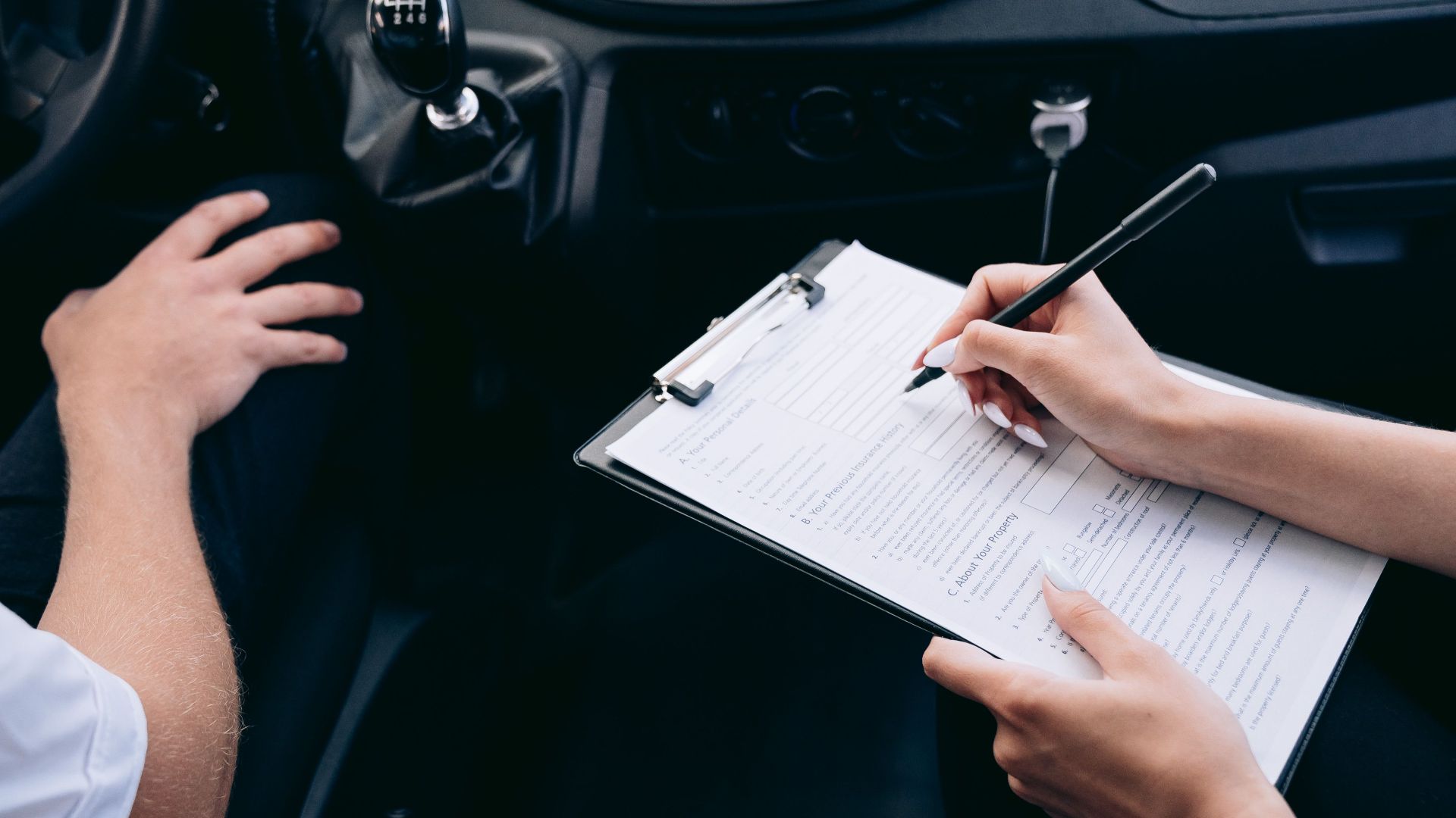 Close-up of a person writing on a clipboard inside a car, showing hands and a gear shift.