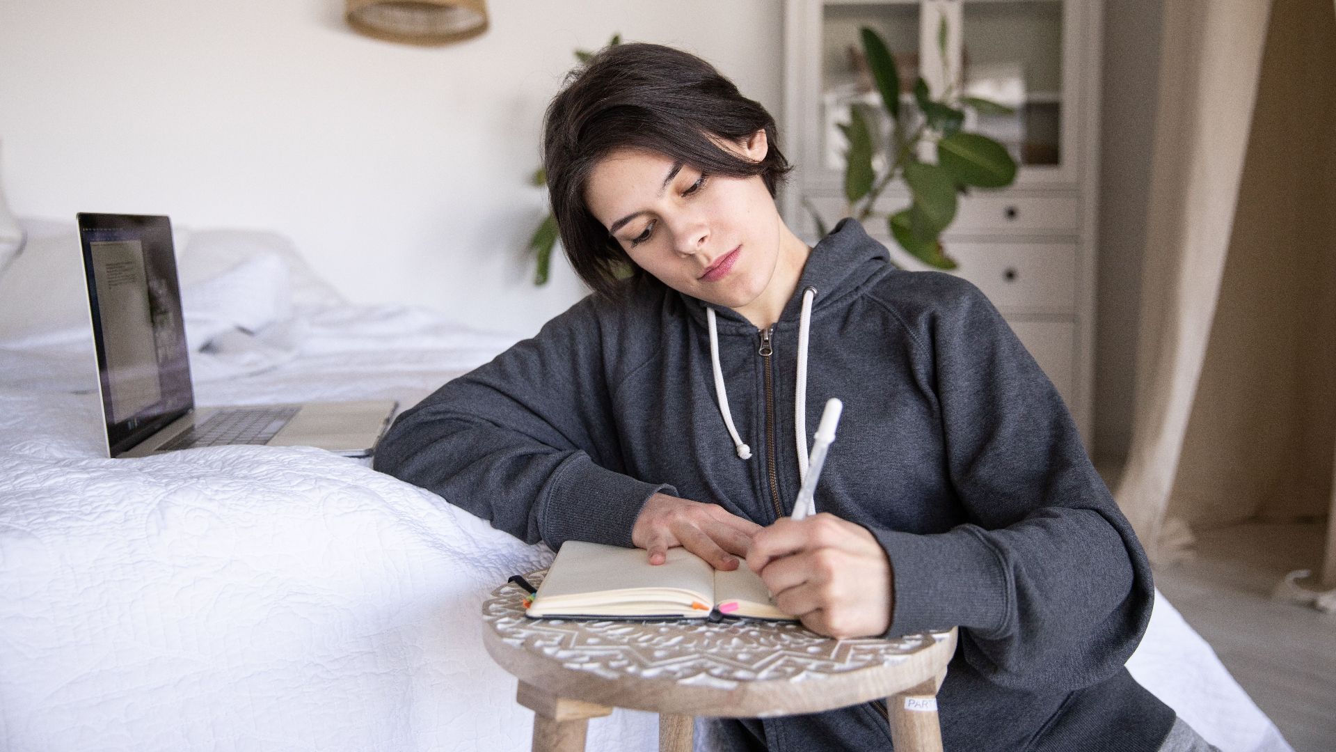 A woman in a hoodie writing notes in a notebook while sitting on a bed in a bright bedroom with a laptop nearby.