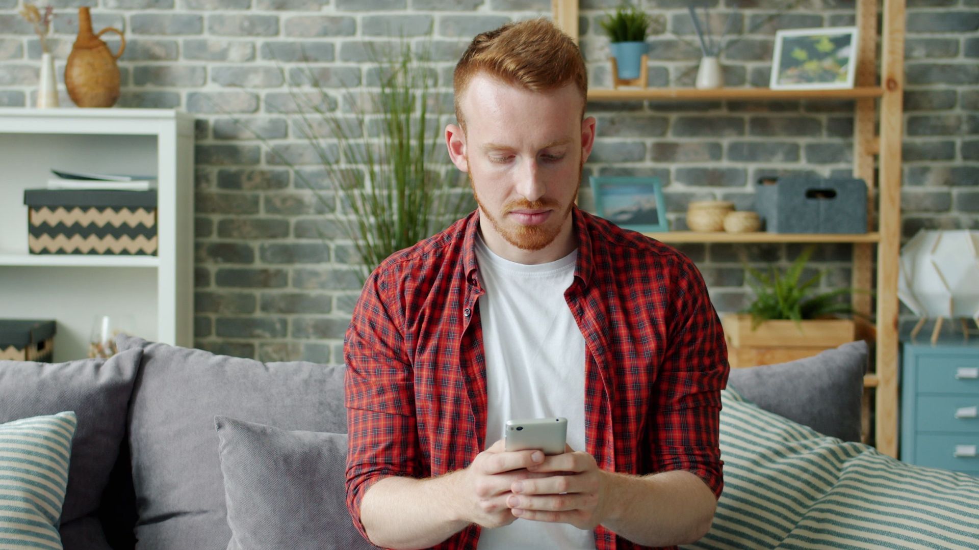 Young man in casual attire sitting on sofa using a smartphone indoors.