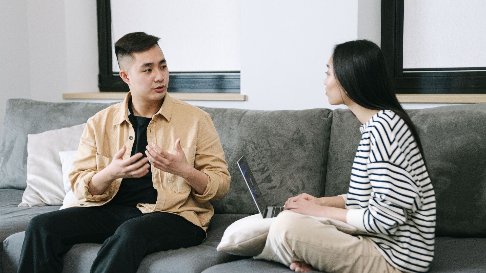 Two young adults engaged in a conversation, sitting on a couch at home.