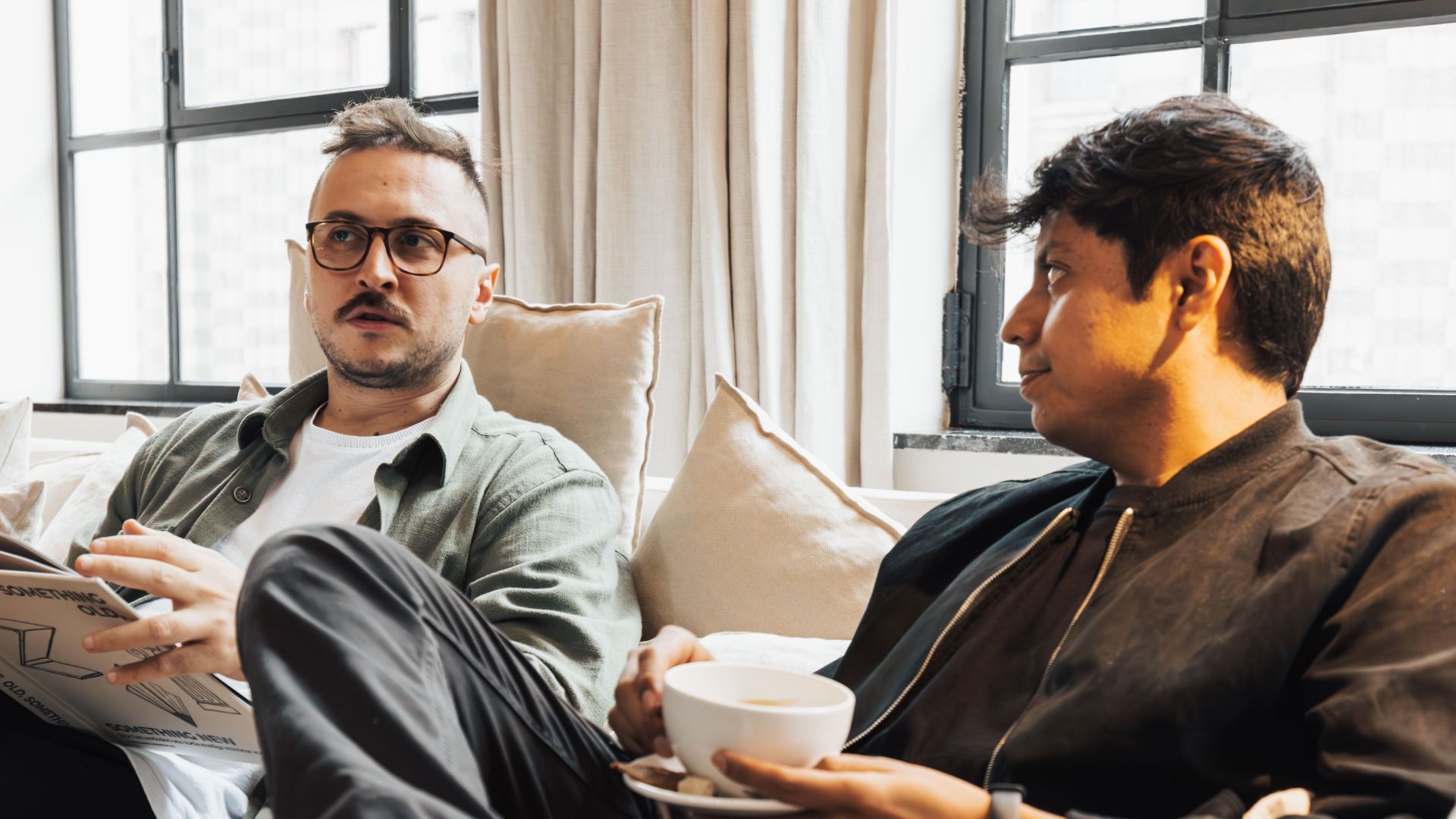 Two men sitting on a cozy sofa, one reading a magazine, enjoying coffee in a bright living room.