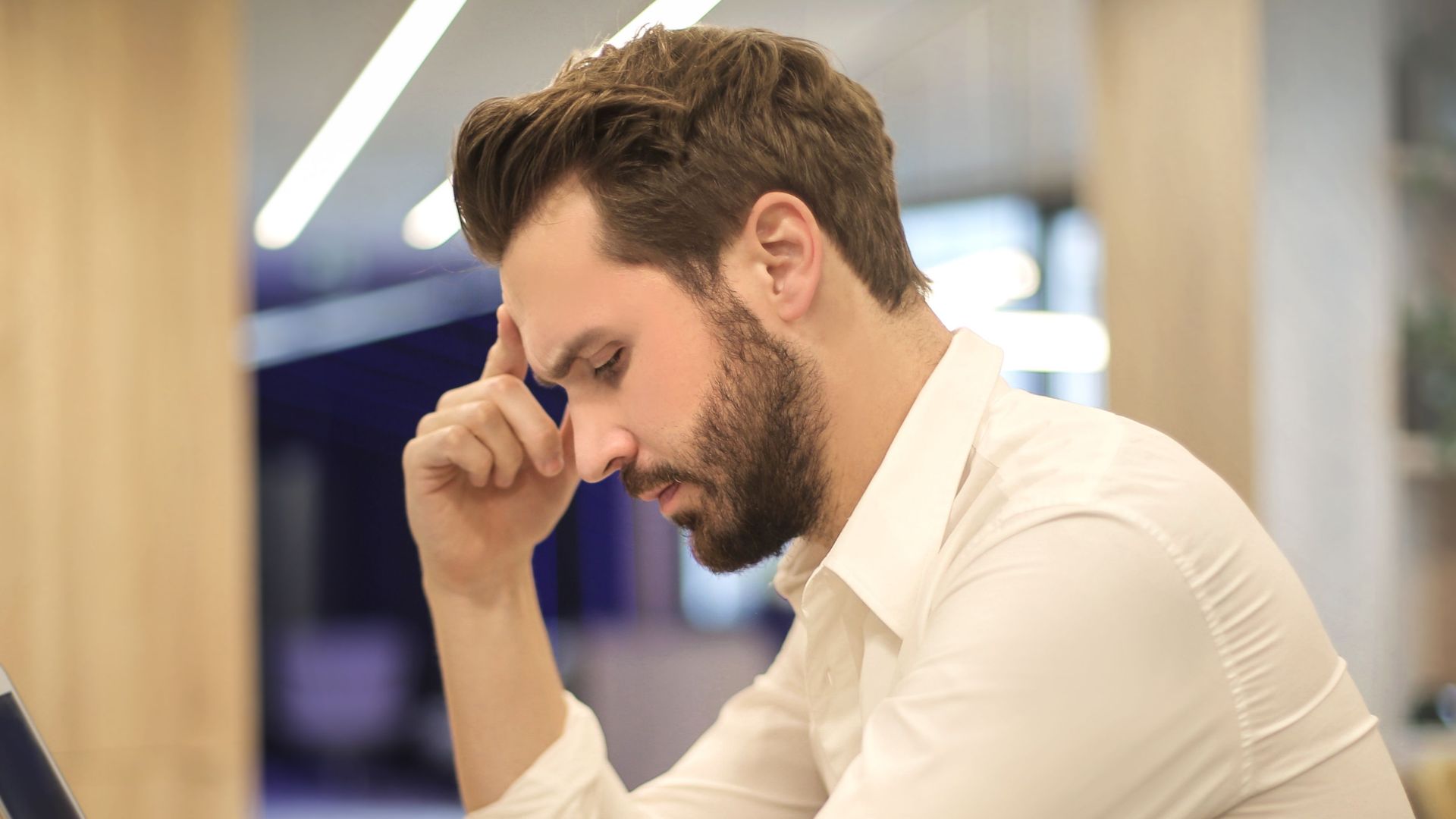 A thoughtful man in a shirt works on his laptop at a modern indoor office space.