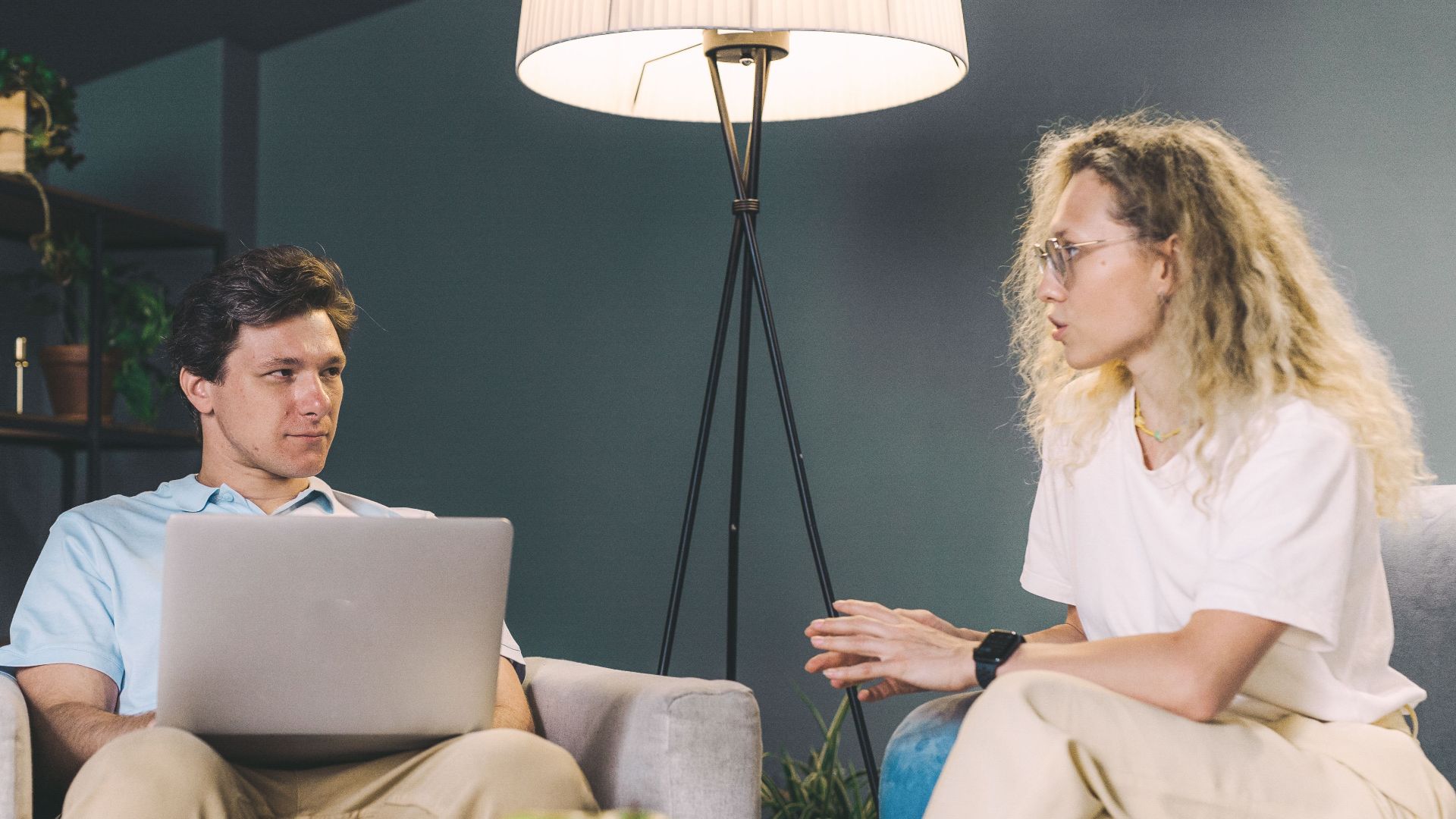 Two adults discuss work while sitting on sofas in a cozy, modern living room setting.