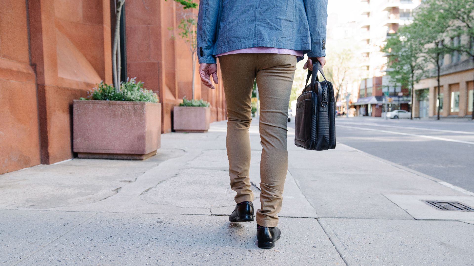 Rear view of a person walking on a city sidewalk holding a briefcase, embodying urban lifestyle.