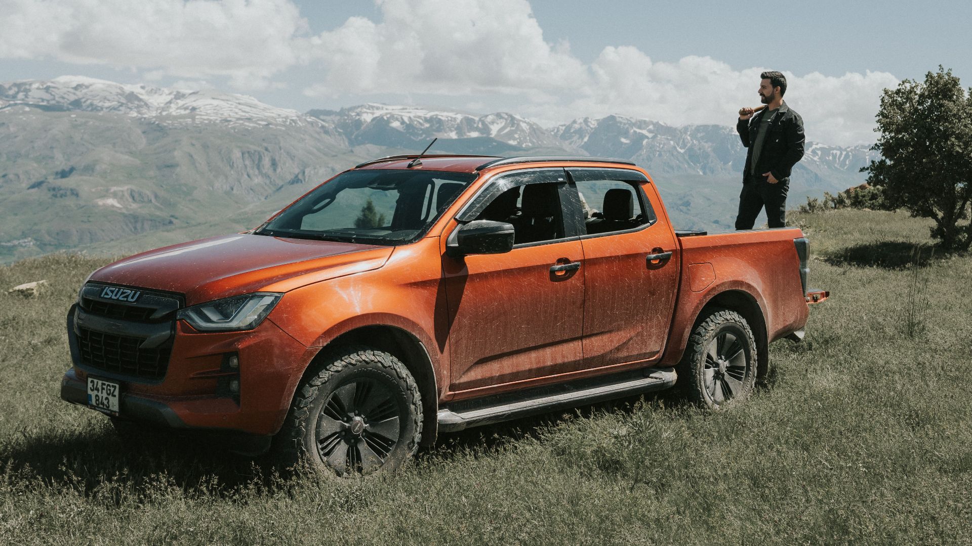 Man with orange pickup truck enjoys picturesque mountain view under a sunny blue sky.