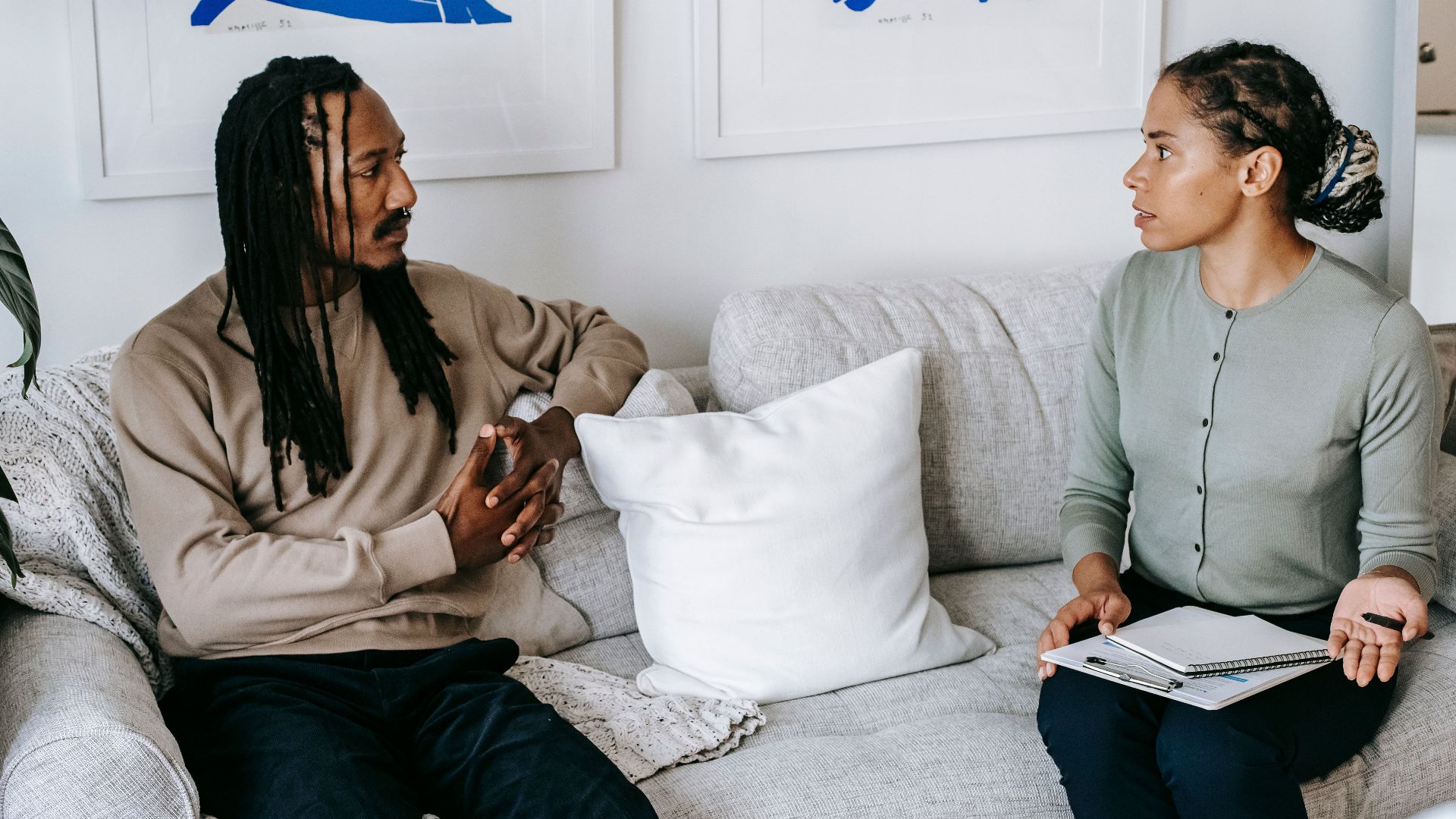 Distressed African American male in smart casual clothes sitting on comfy sofa and listening to professional female counselor in psychology center