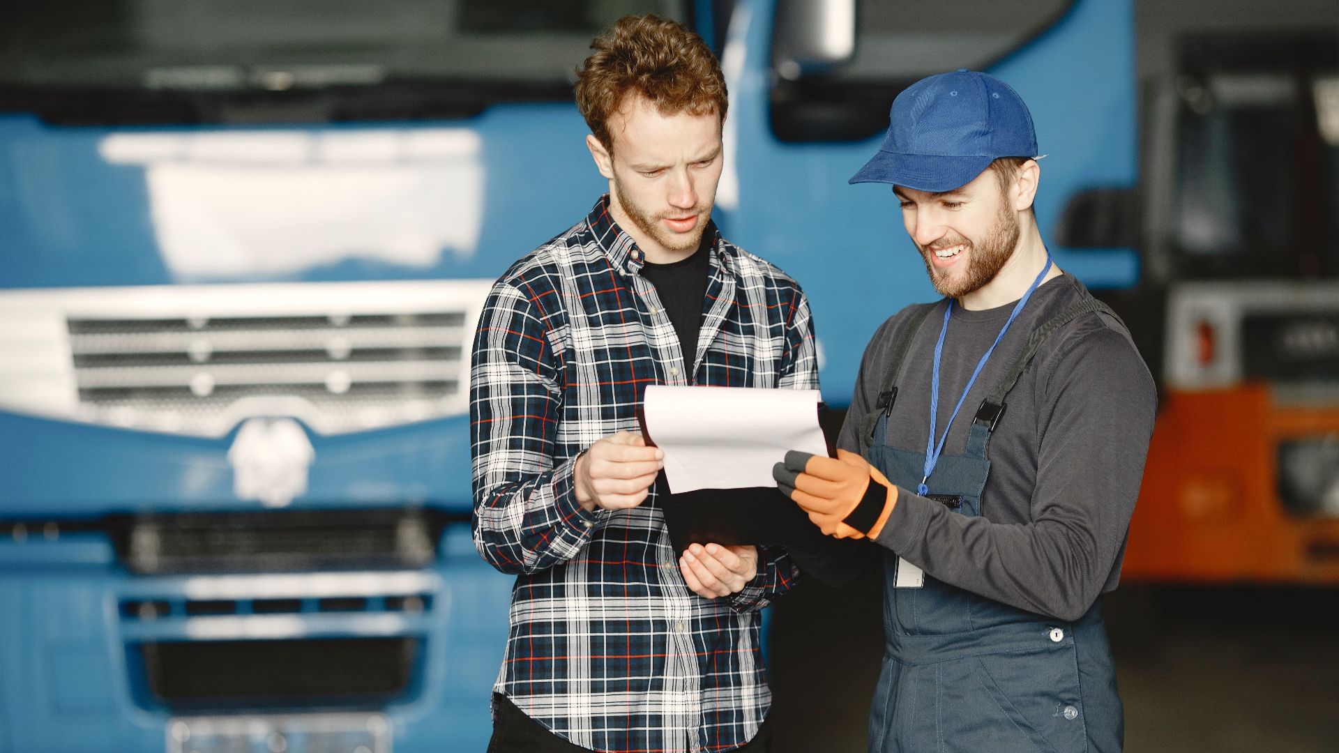 Two men in discussion over vehicle maintenance documents in a garage setting with trucks in the background.