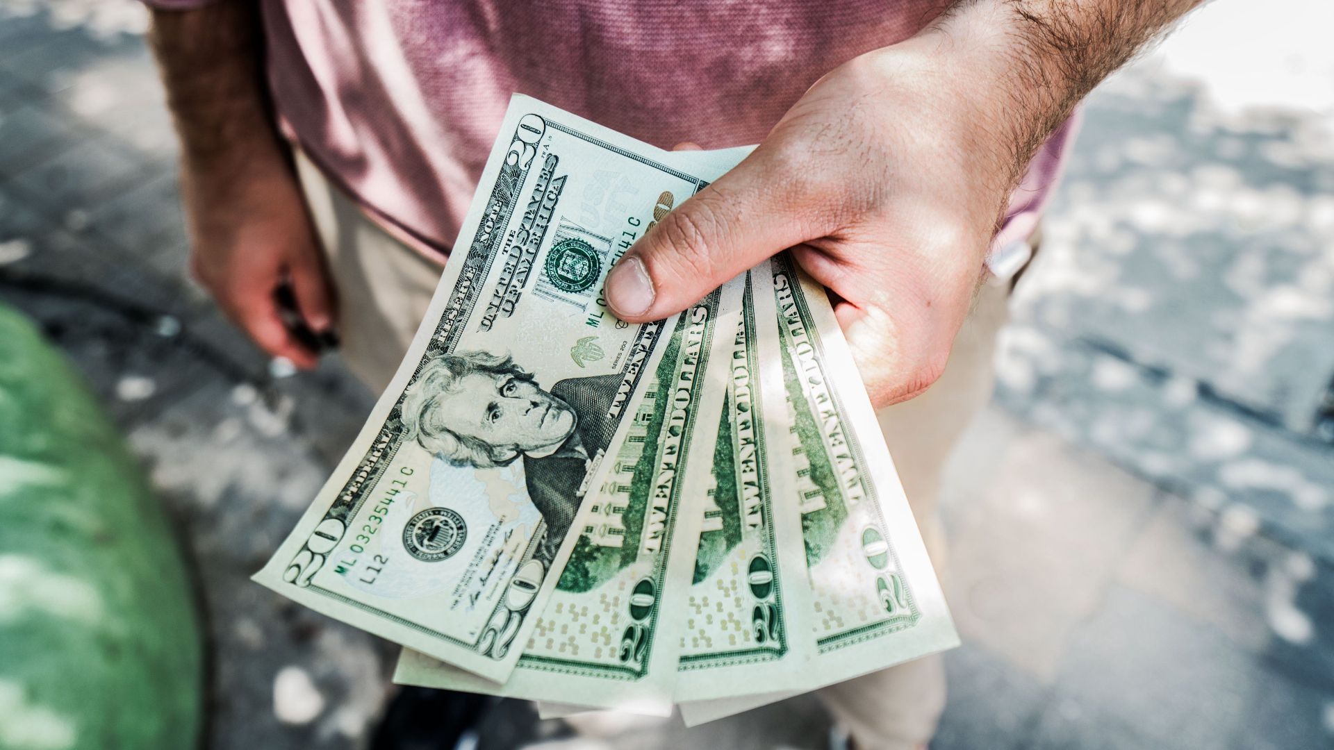 A person holding several twenty-dollar bills outdoors, depicting payment or financial transaction.