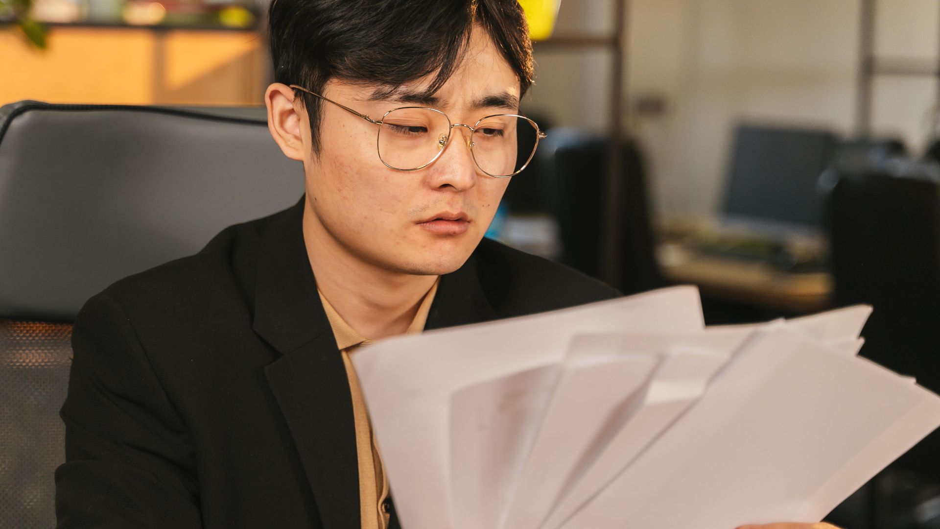 Asian man in glasses and suit analyzing papers at his desk in a modern office.