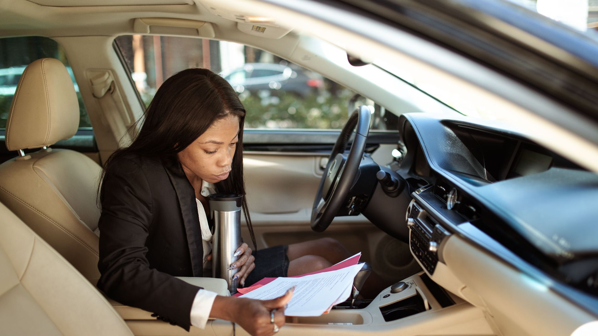 A focused businesswoman reads documents in her car, multitasking with a tumbler.