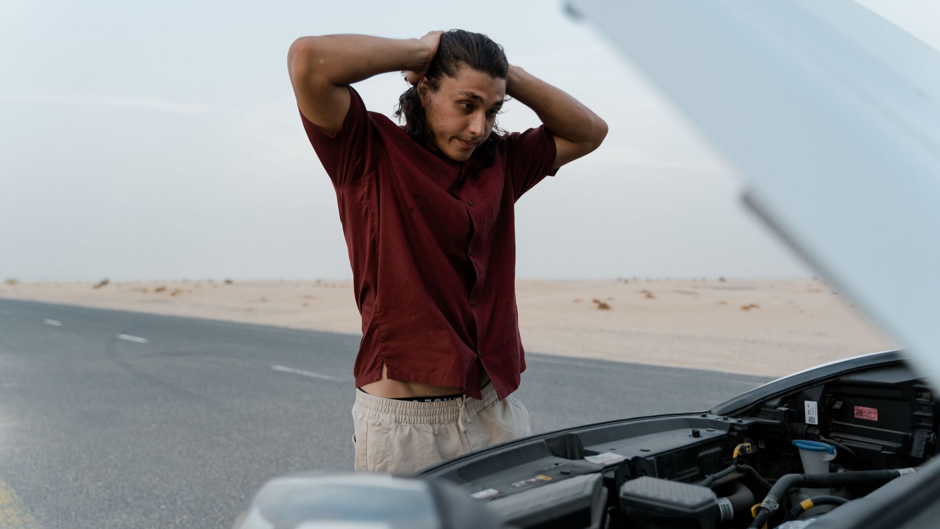 A frustrated man examining a broken car engine on a deserted road, showing stress and concern.