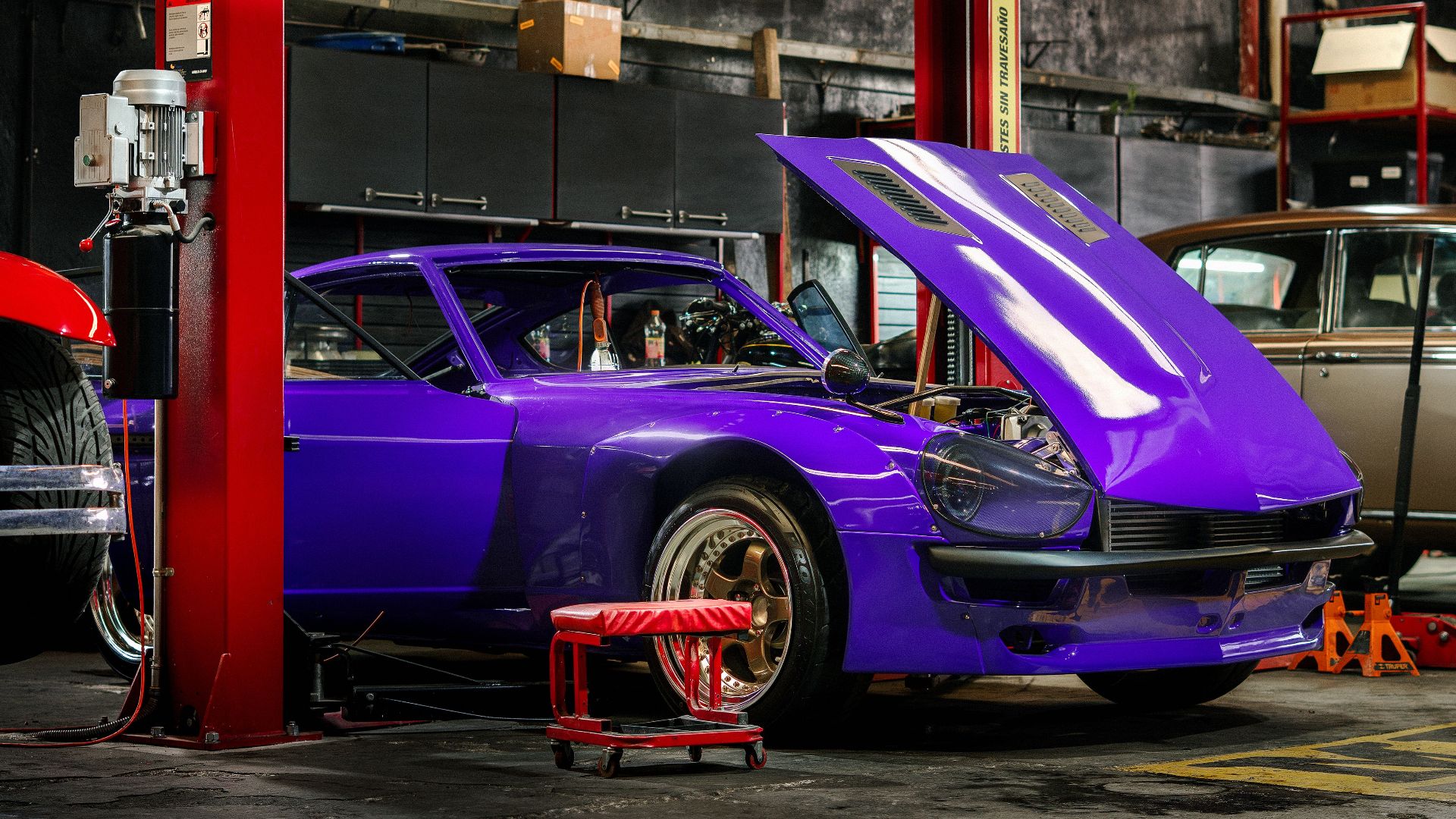 A classic car with a vibrant purple color in a mechanical workshop with its hood open.