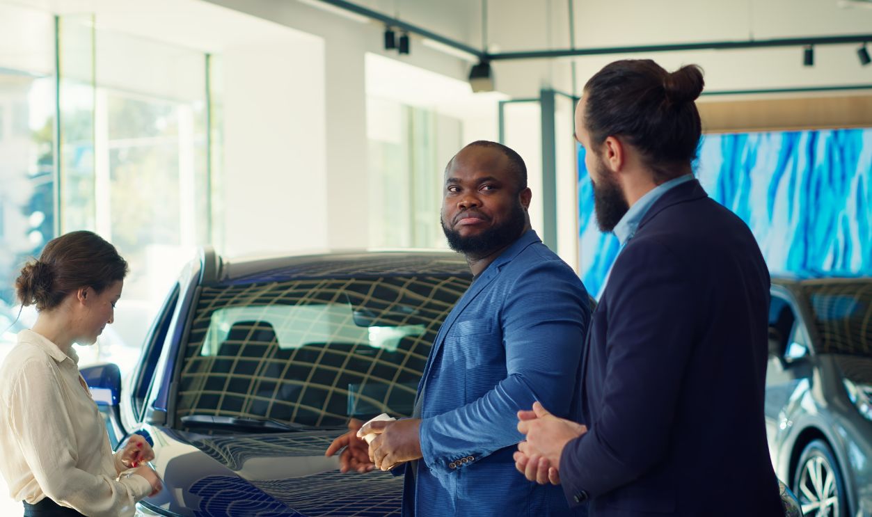 Clients arriving in car dealership, being greeted by friendly car salesman ready to showcase available vehicles to clients. Customers walking in showroom, being welcomed by agent, camera A