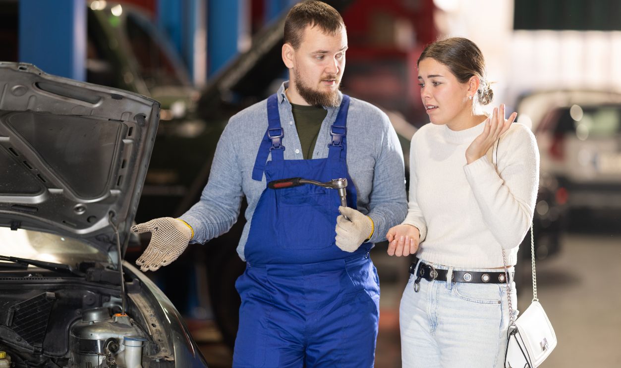 Male mechanic advises young woman client on repairing under hood of car in car service station