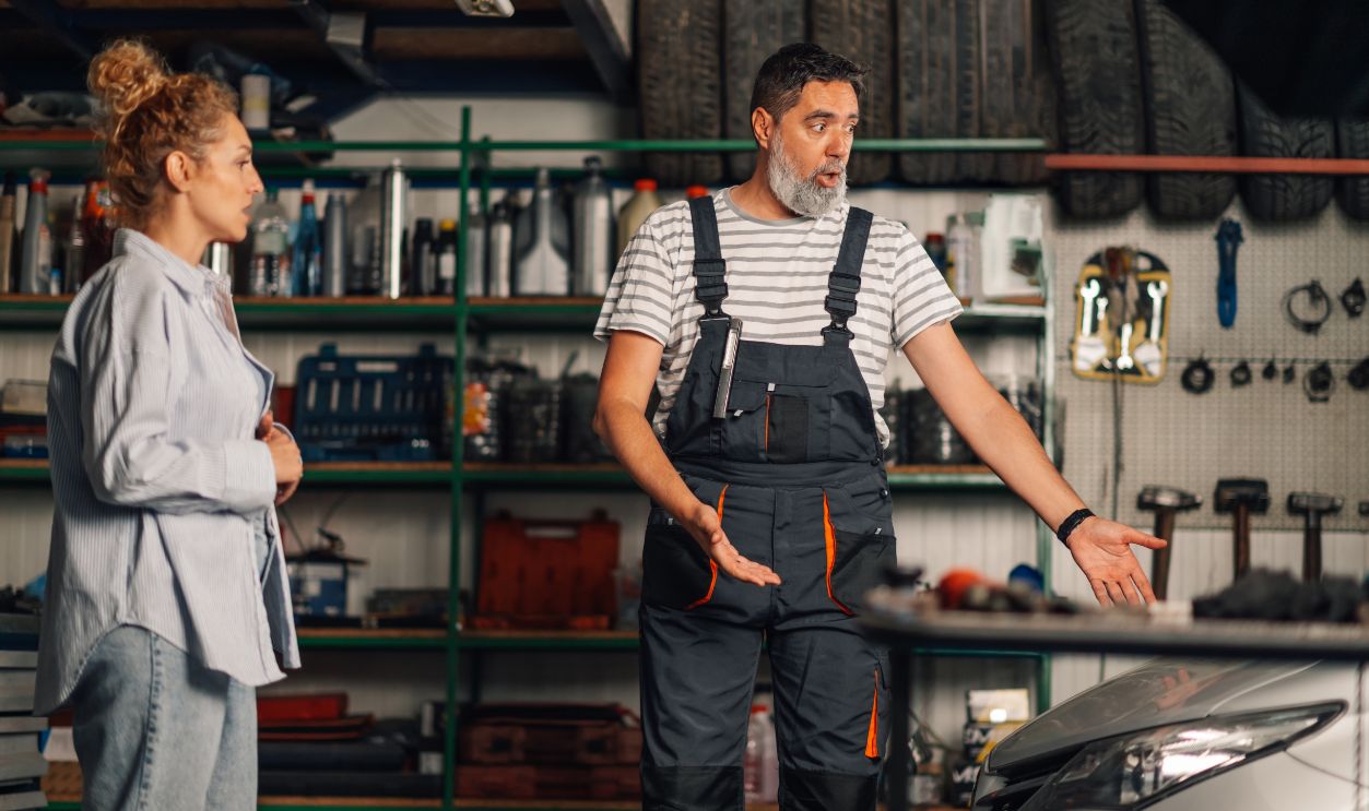 Nervous serviceman in uniform is standing at auto mechanic workshop near broken car and looking at it while yelling at confused woman. Angry auto mechanic is explaining customer damage she made.