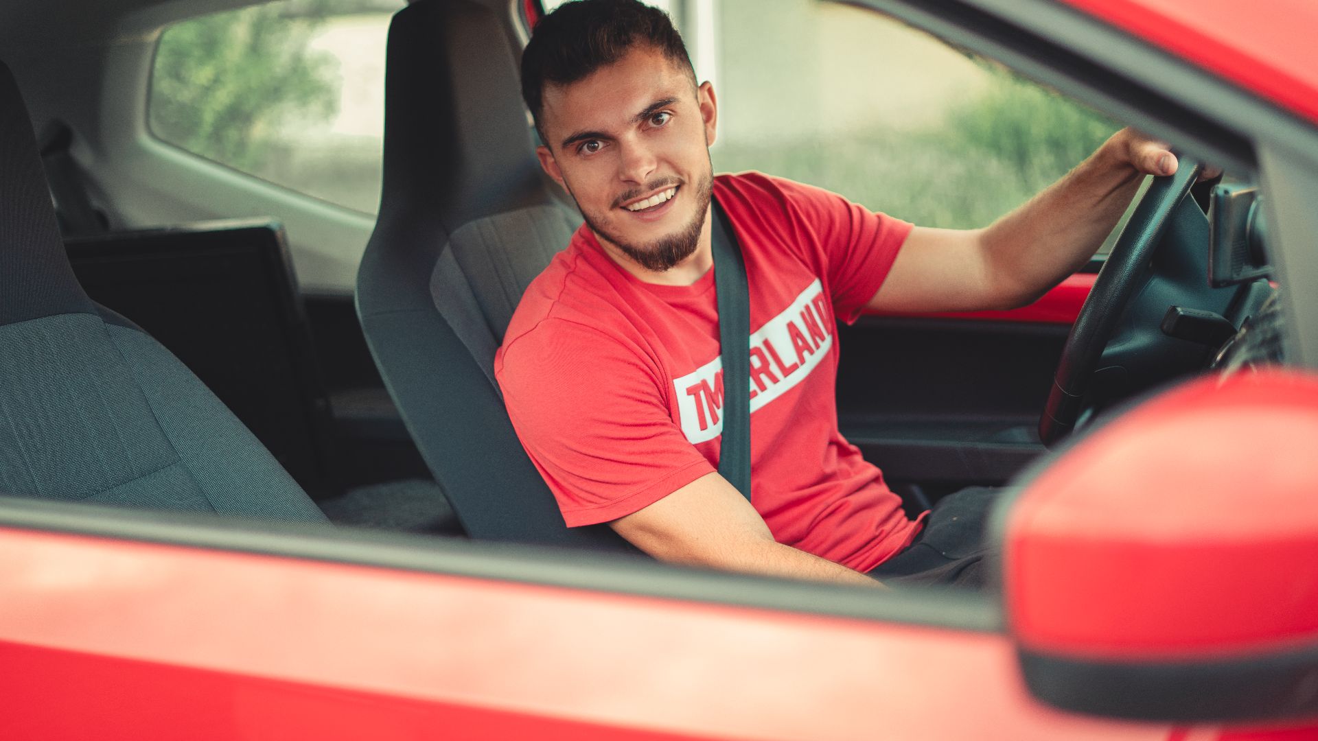 Smiling man driving a red car while wearing a seatbelt, showcasing safety and joy.
