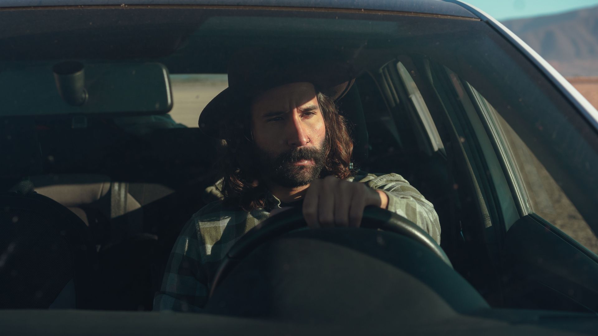 A bearded man in a hat driving a car through a sunny desert landscape, conveying a sense of adventure.
