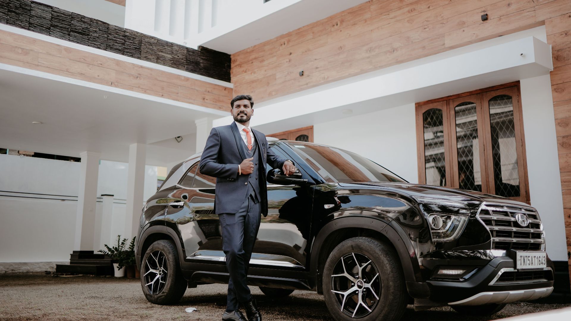 Businessman in a blue suit standing confidently next to a modern black SUV outside a stylish home in India.