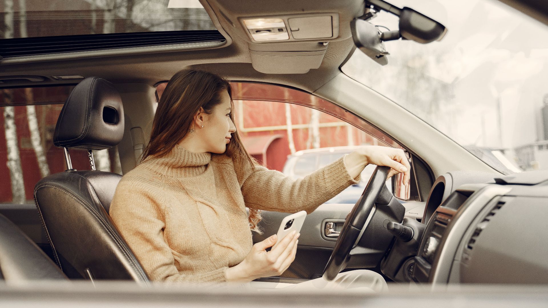 From window side view of adult woman in warm clothes sitting at wheel of modern car waiting for friend and using smartphone
