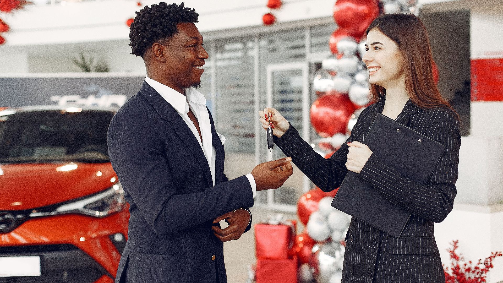 Side view of happy positive female dealer in formal classy wear passing car keys to  excited African American customer in trendy suit in car showroom