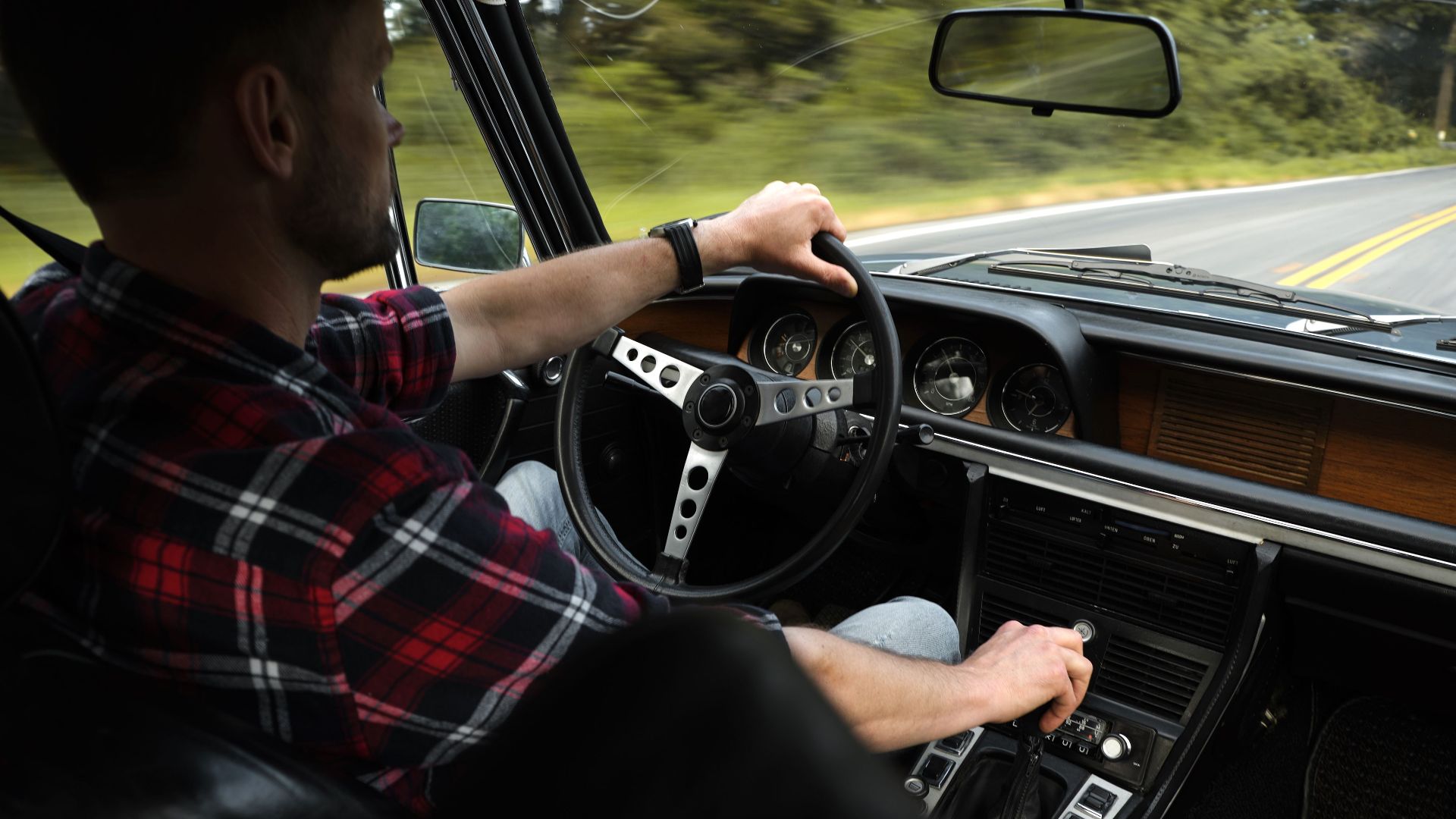 Back view of serious unshaven man in plaid shirt driving automobile by asphalt road among green trees