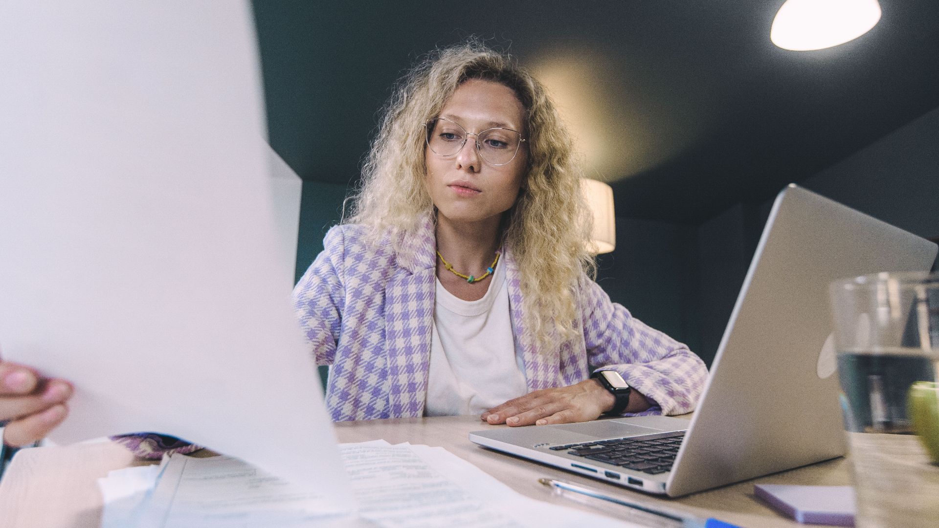 Woman in an office environment reviewing documents with focus, surrounded by technology.