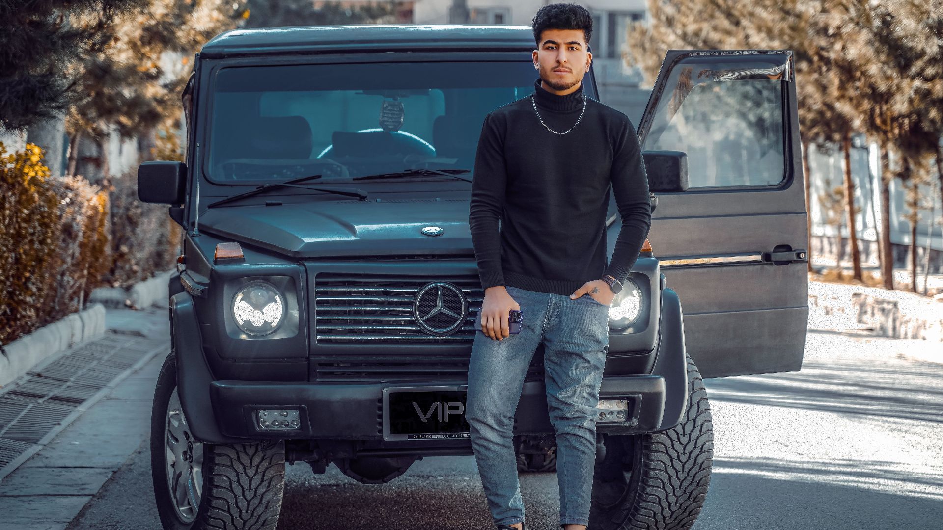 A fashionable young man stands confidently in front of a black Mercedes SUV, showcasing style and luxury.