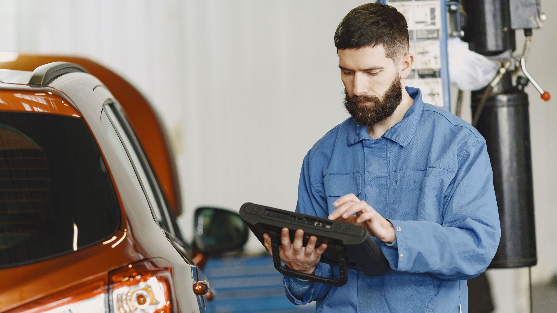 A mechanic in coveralls using a diagnostic tool at an auto repair shop beside a car.