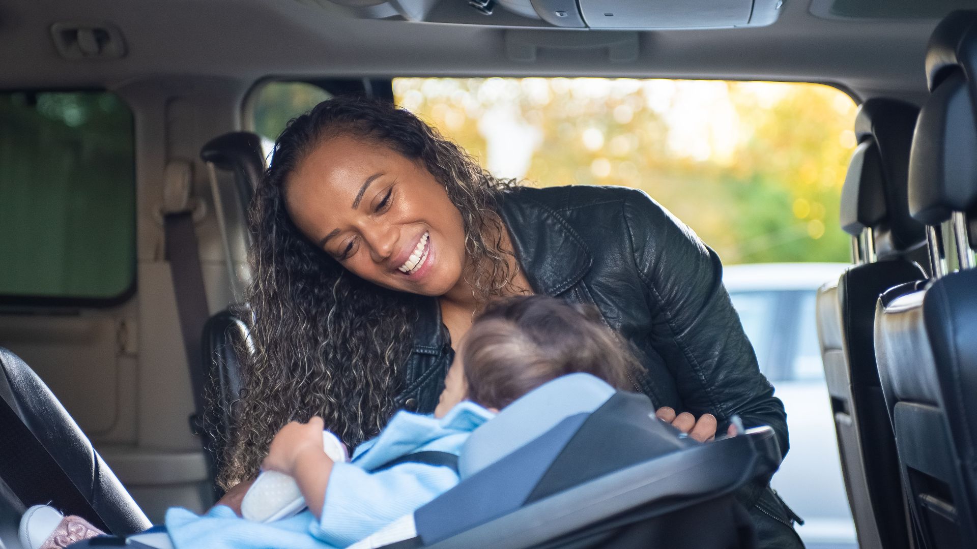 A joyful mother smiles at her baby in a car seat inside a vehicle.