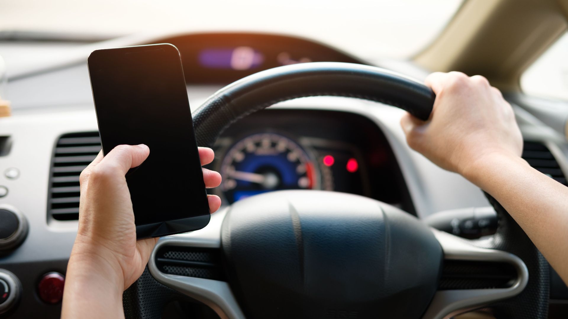 A driver uses a smartphone while steering a vehicle, showcasing distracted driving risks.